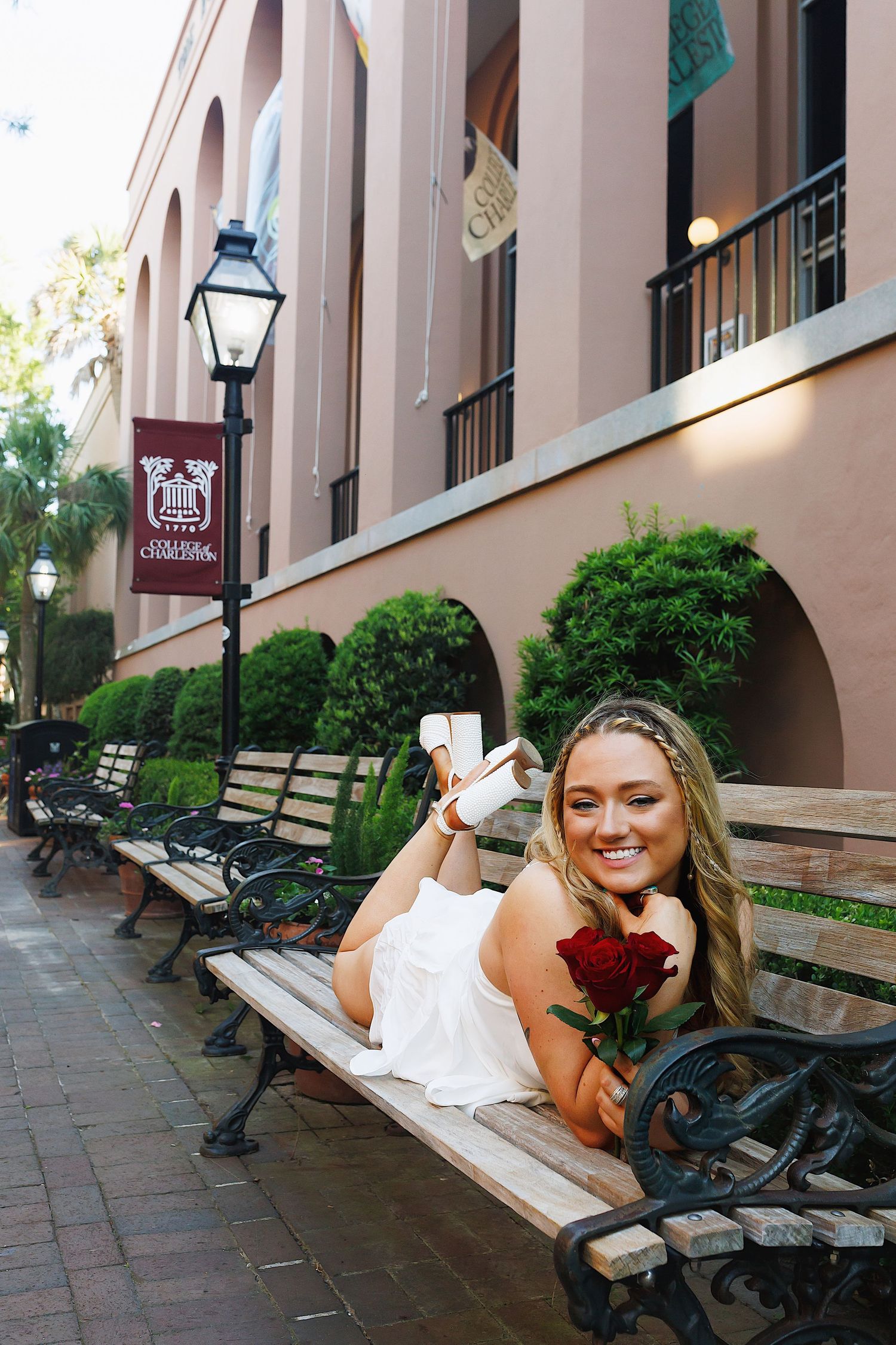 A person in a white dress poses on a park bench along a brick walkway with arched architecture in the background.