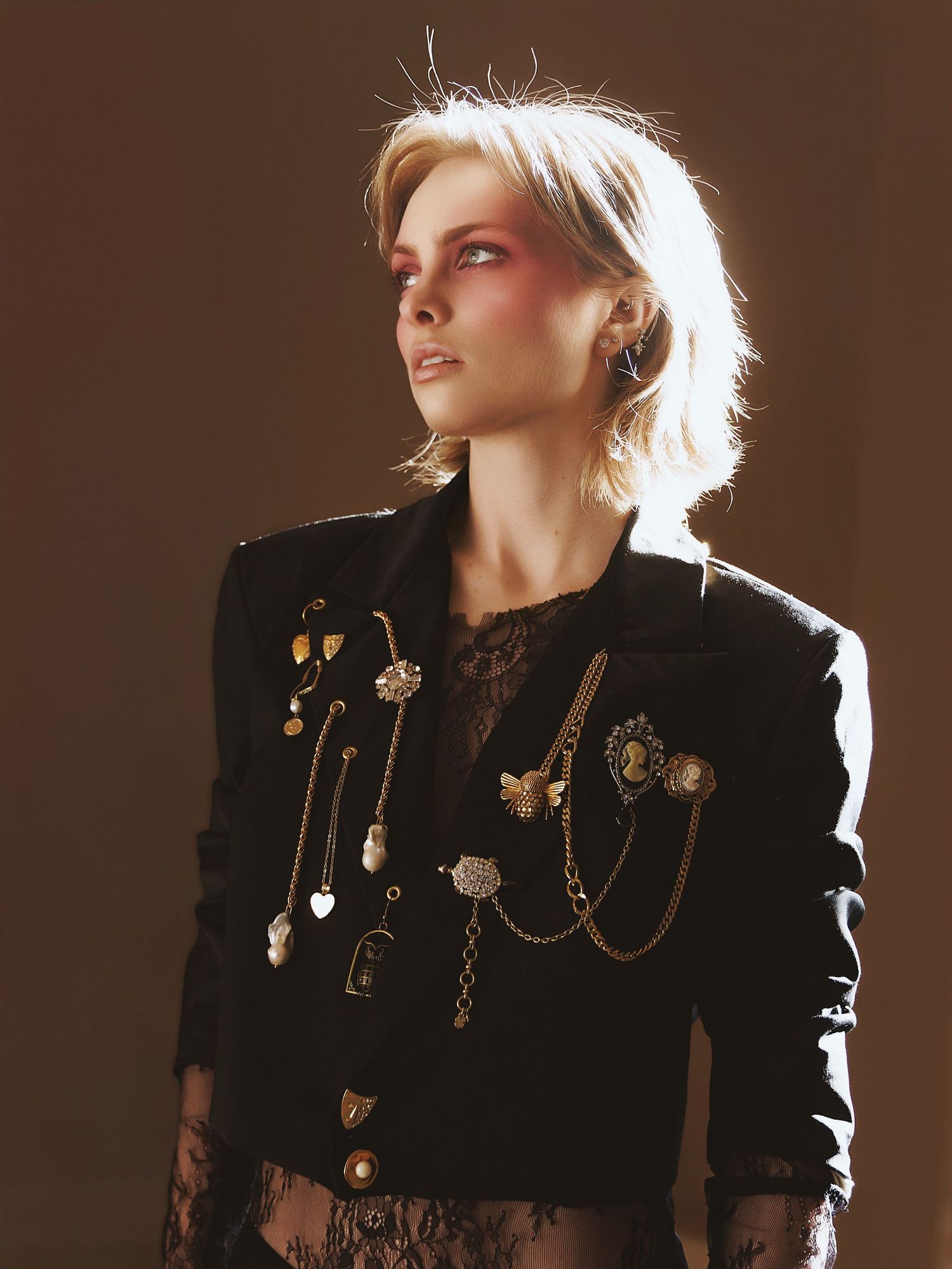 Fashion portrait showing ornate vintage-style brooches and chains adorning a black blazer against warm brown lighting.