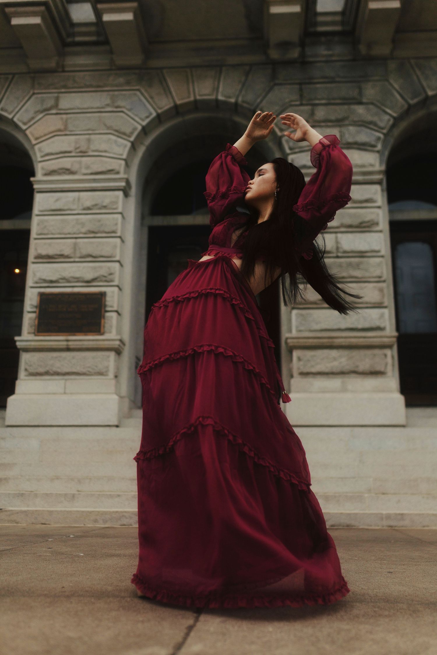 A person in a flowing burgundy tiered dress strikes a dramatic pose against a classical stone building facade.