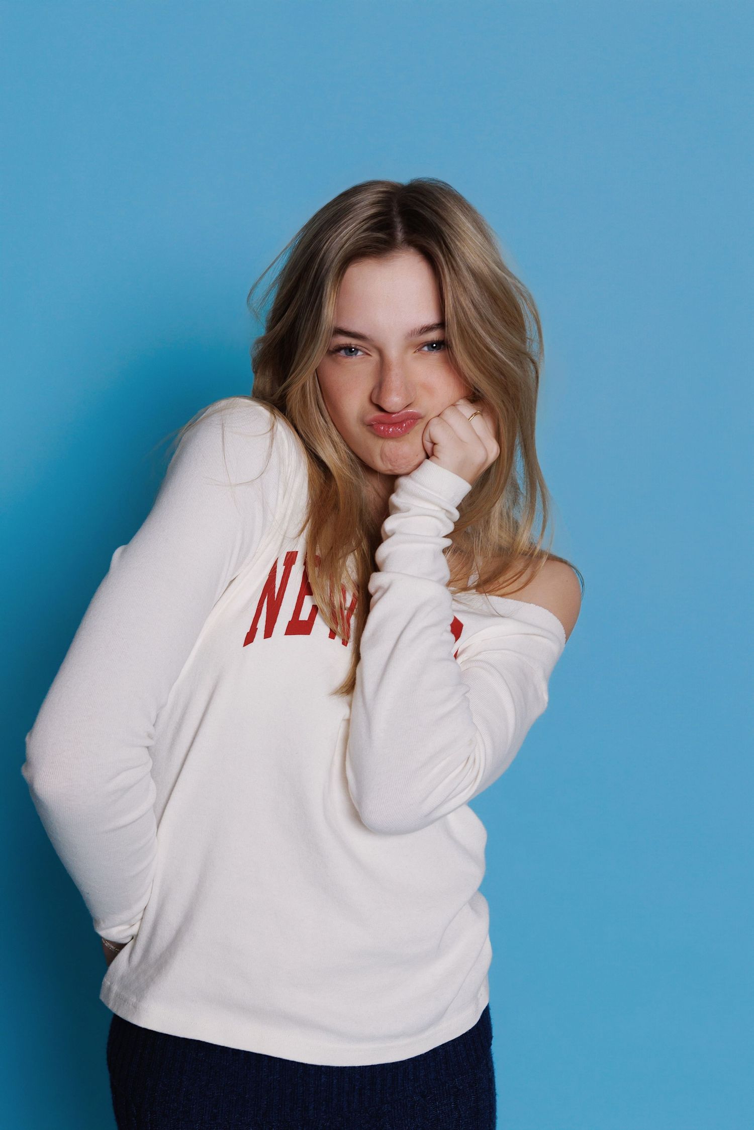 A person in a white off-shoulder sweater poses playfully against a bright blue background.