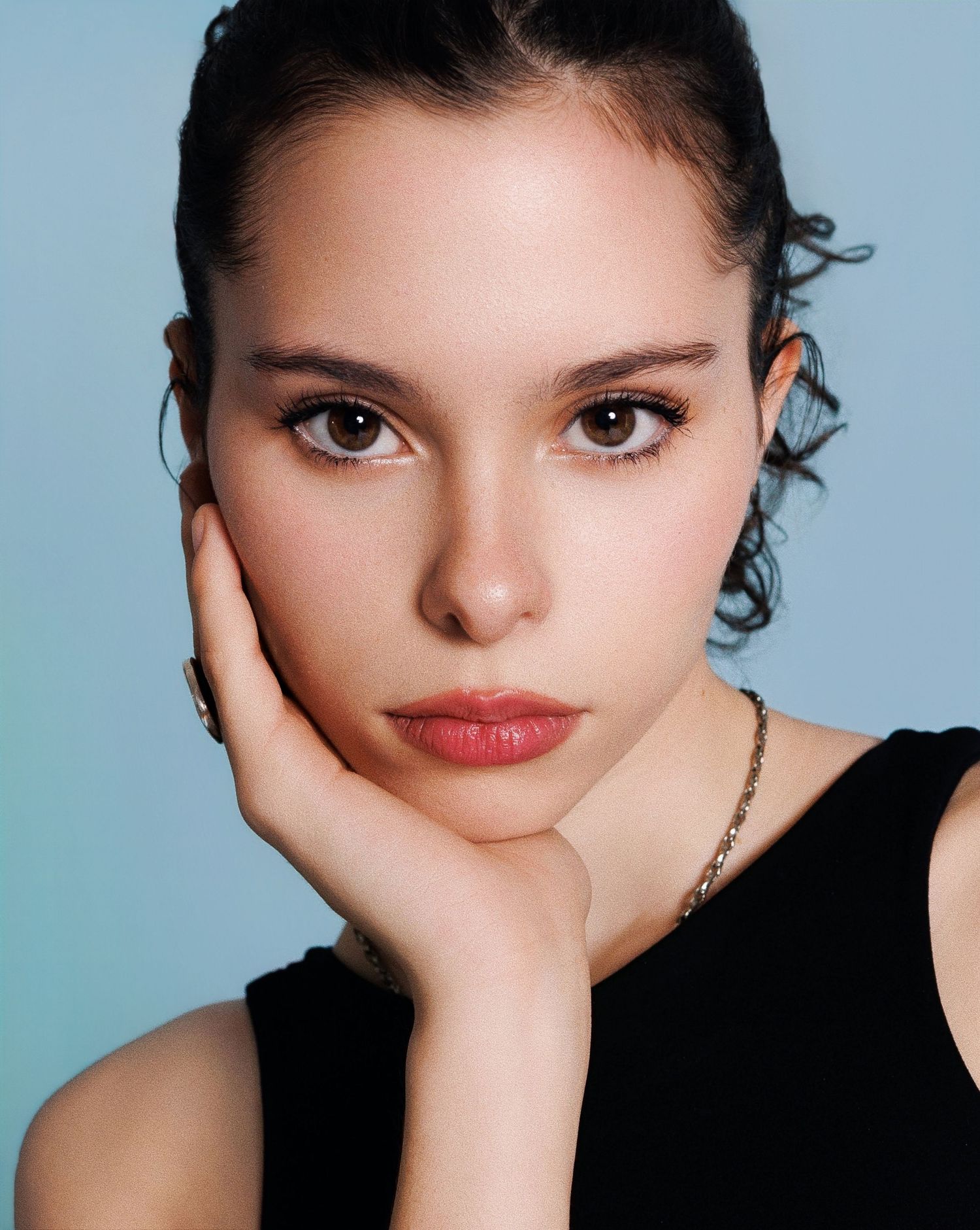 Portrait of someone with dark hair and red lips against light blue background, resting face on hand in black top.