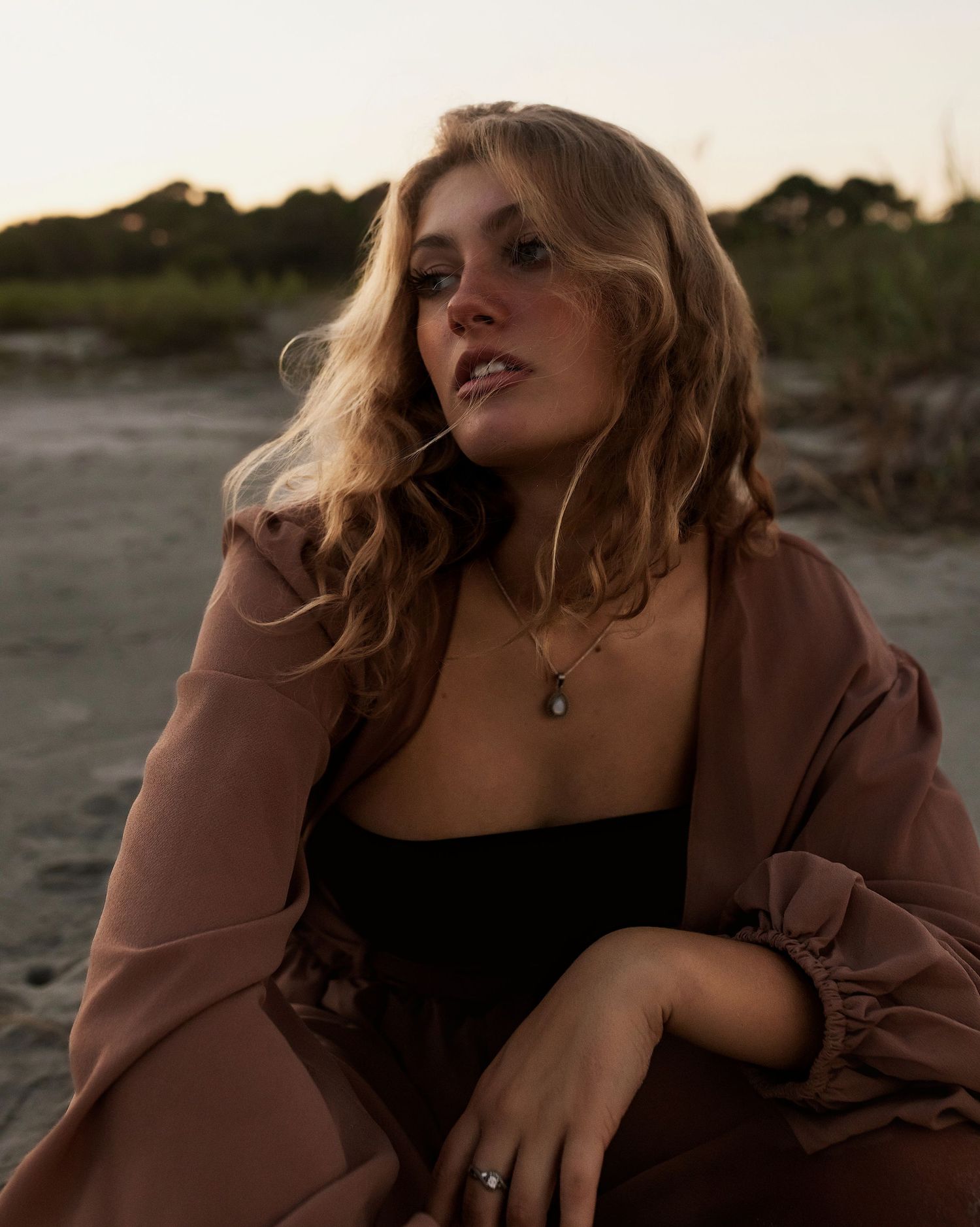 A portrait taken at sunset showing someone in a brown jacket and black top against a natural outdoor backdrop.