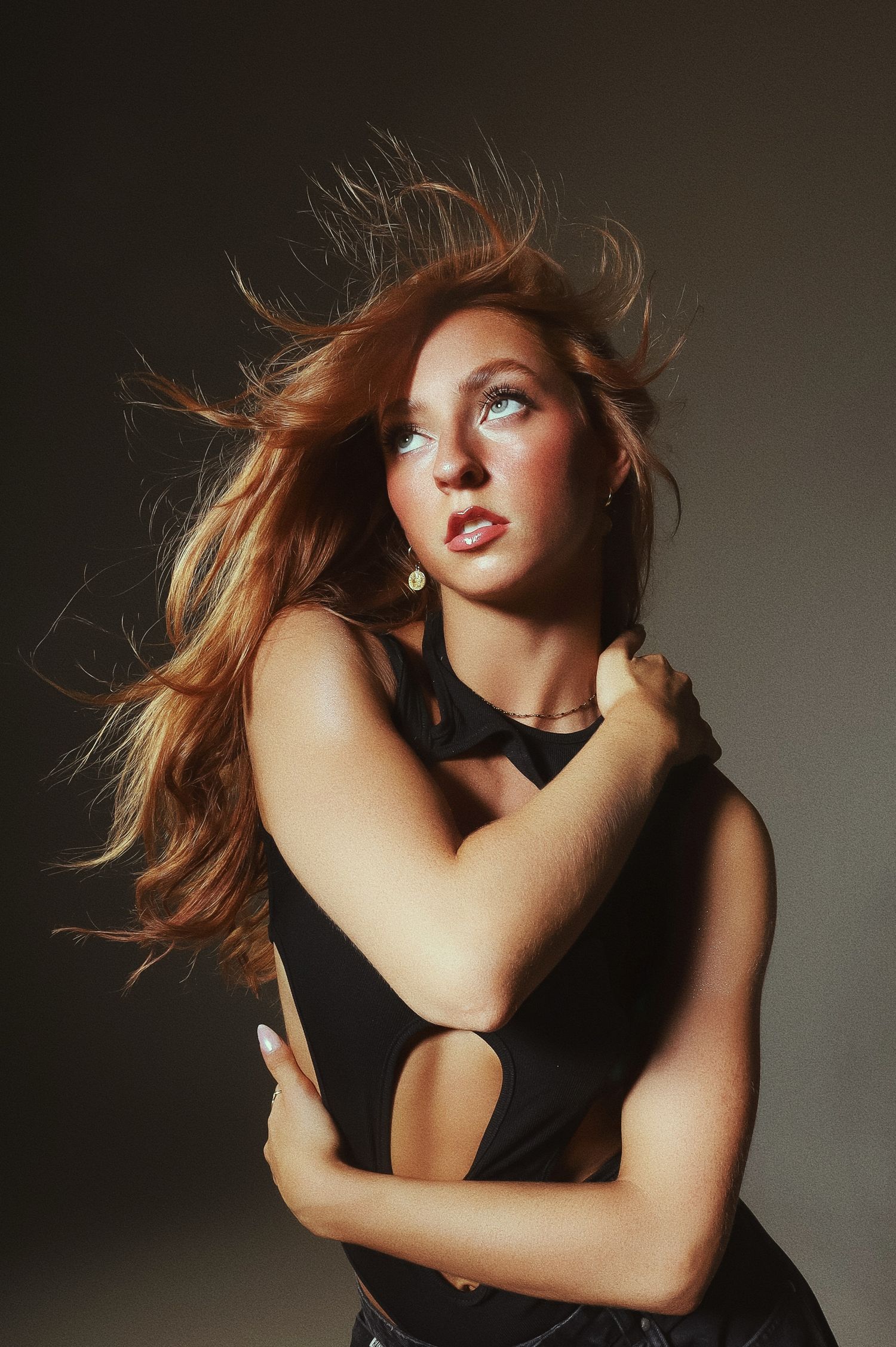 Dramatic fashion portrait with windswept auburn hair and black cutout dress against dark background with studio lighting.
