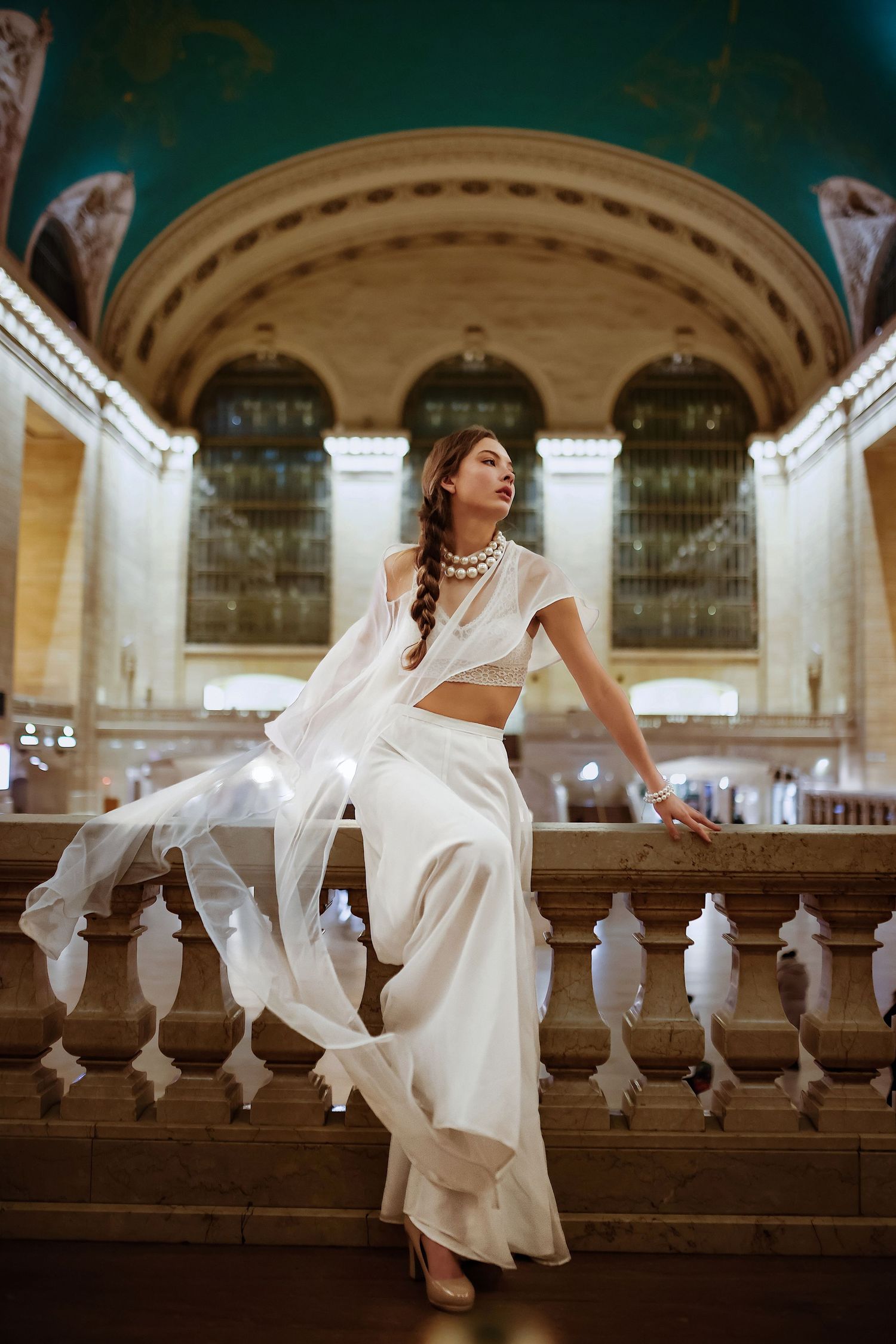 A person in a flowing white dress poses gracefully along a classic stone balustrade in an elegant architectural setting.