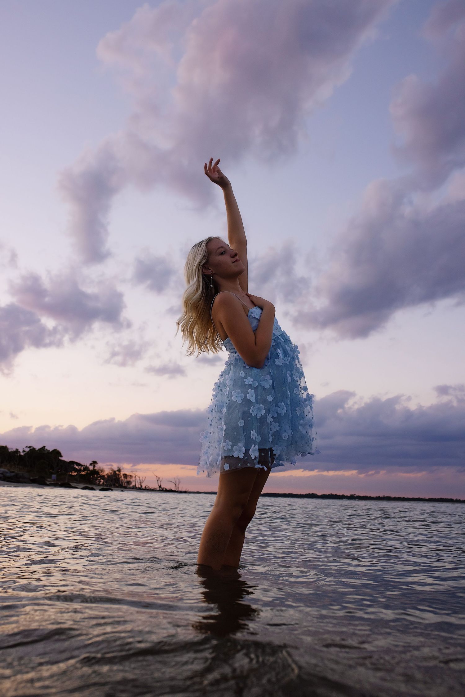 A figure in a blue dress splashes and dances in ocean waters during a dramatic purple sunset.