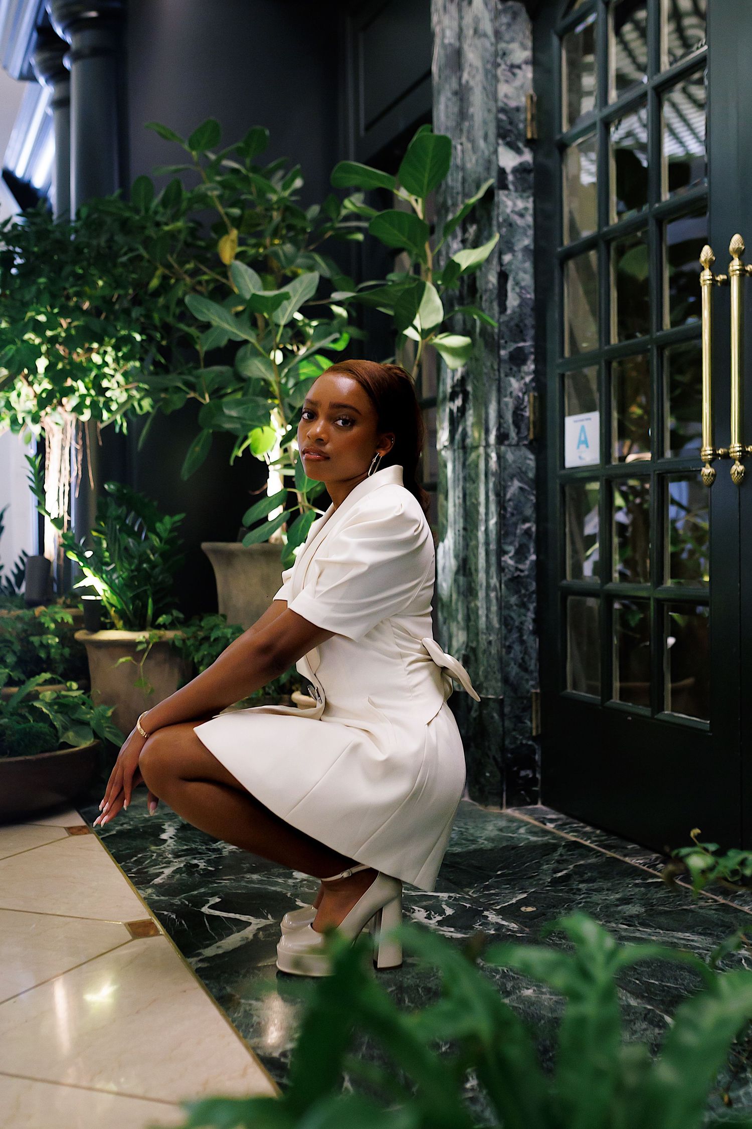Person in white dress sits elegantly among lush indoor plants near glass windows creating a serene greenhouse atmosphere.