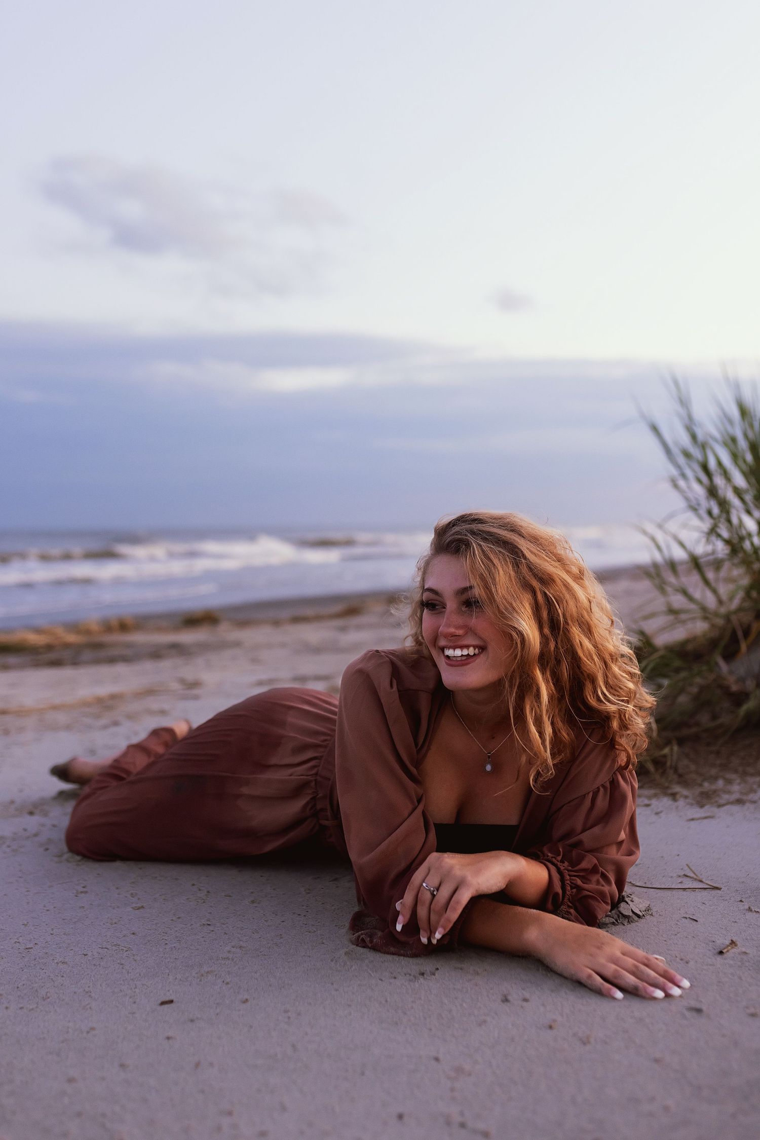 Person in brown sweater lying on beach sand at sunset with crashing waves and cloudy sky in background.