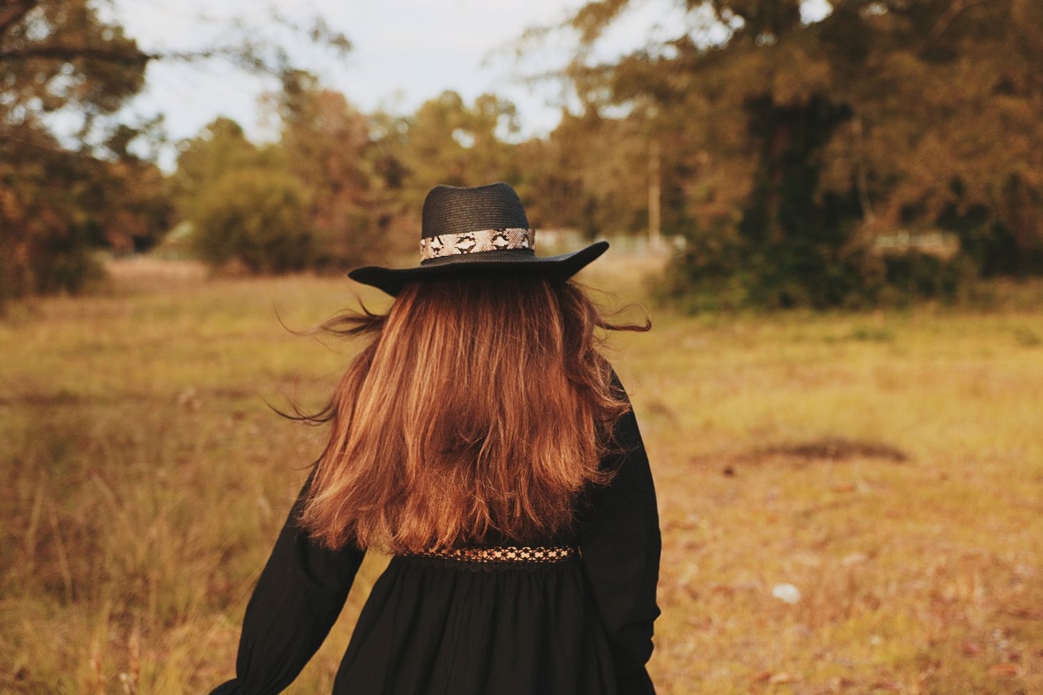 A person with long red hair stands in a golden autumn field at sunset wearing a black outfit and floral crown.