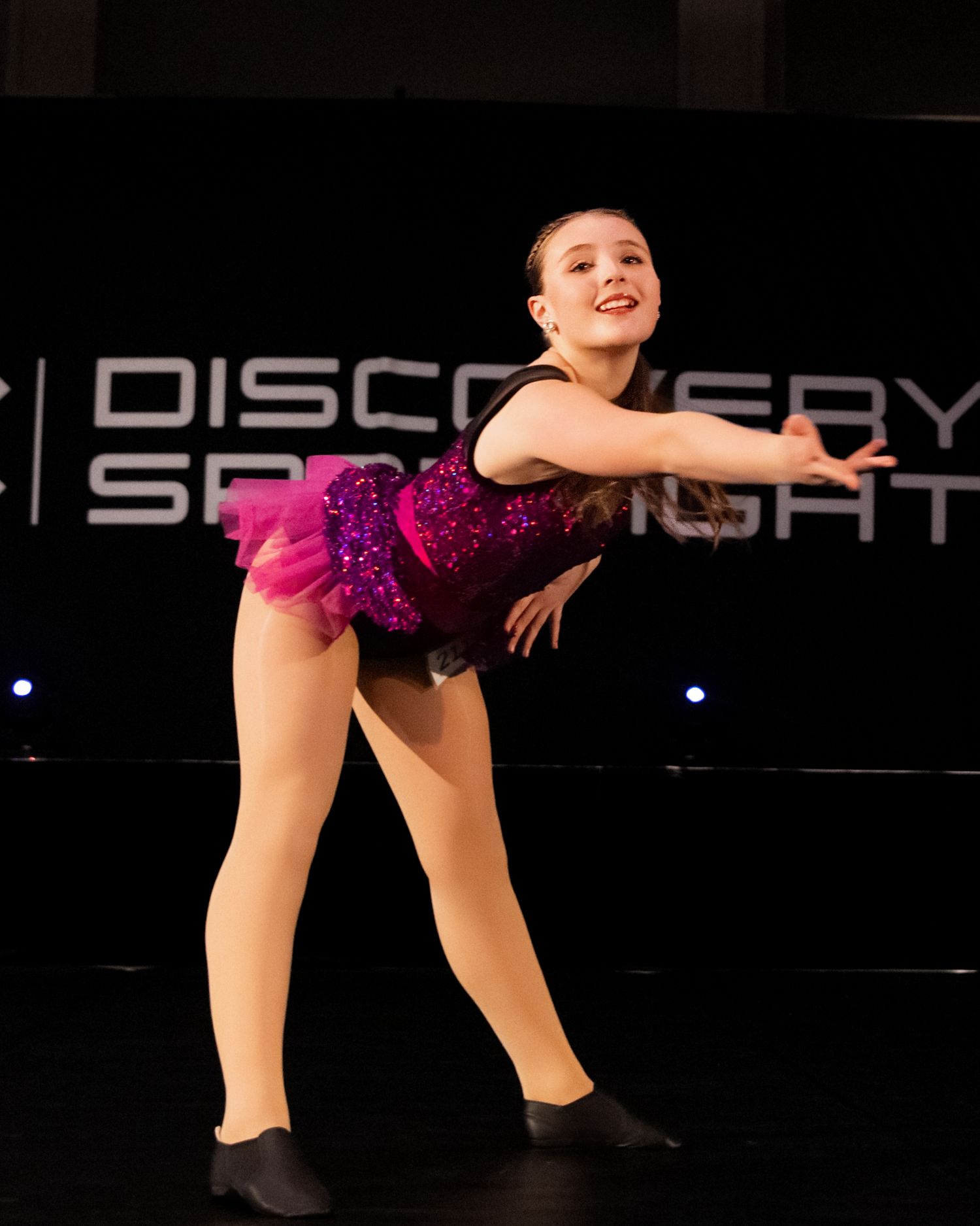 Dancer in black and pink costume performs graceful split leap on dark stage with illuminated Discovery sign in background.