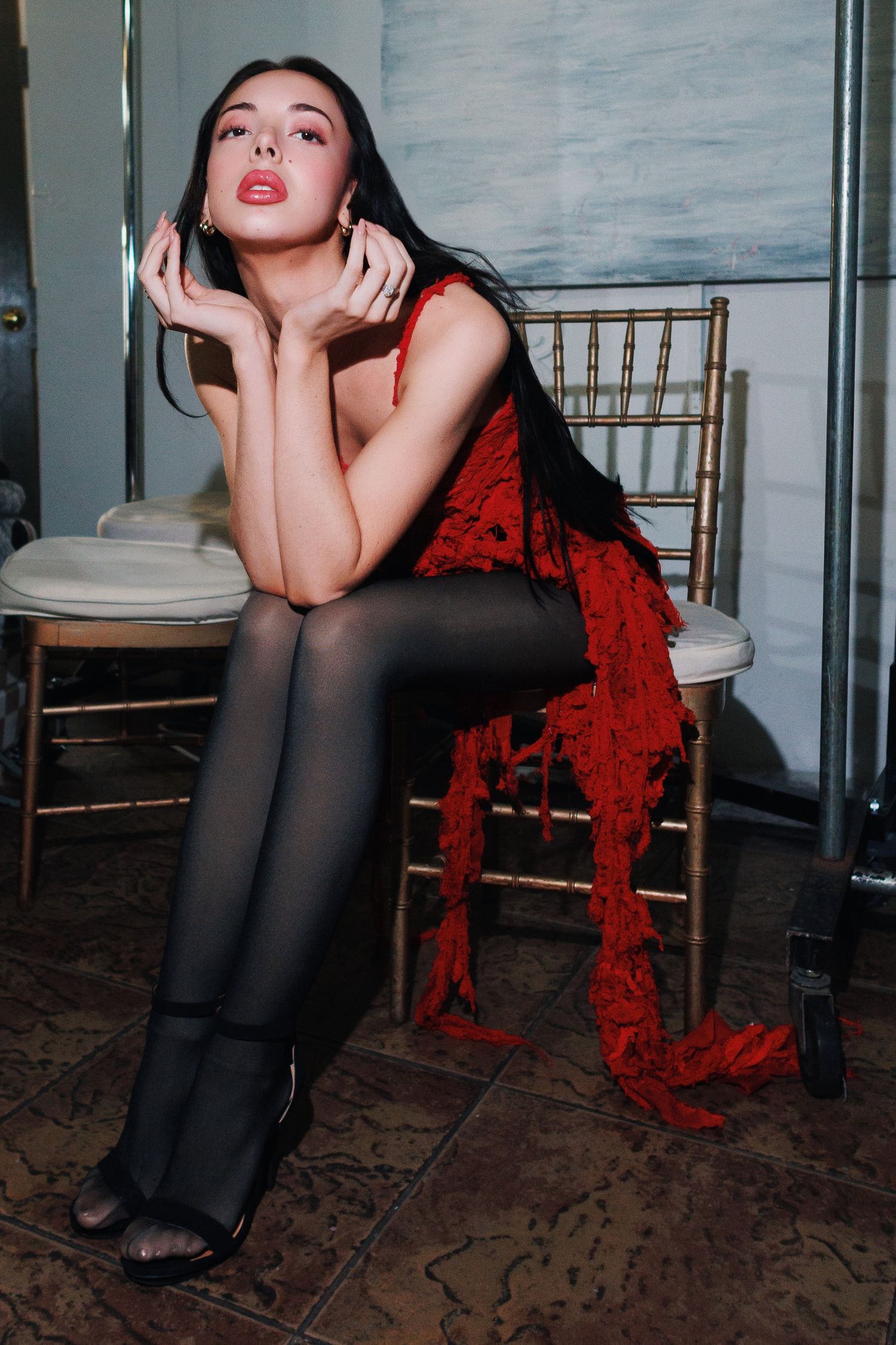 Fashion model in black stockings sits on gold chair with flowing red fabric in artistic photo series.