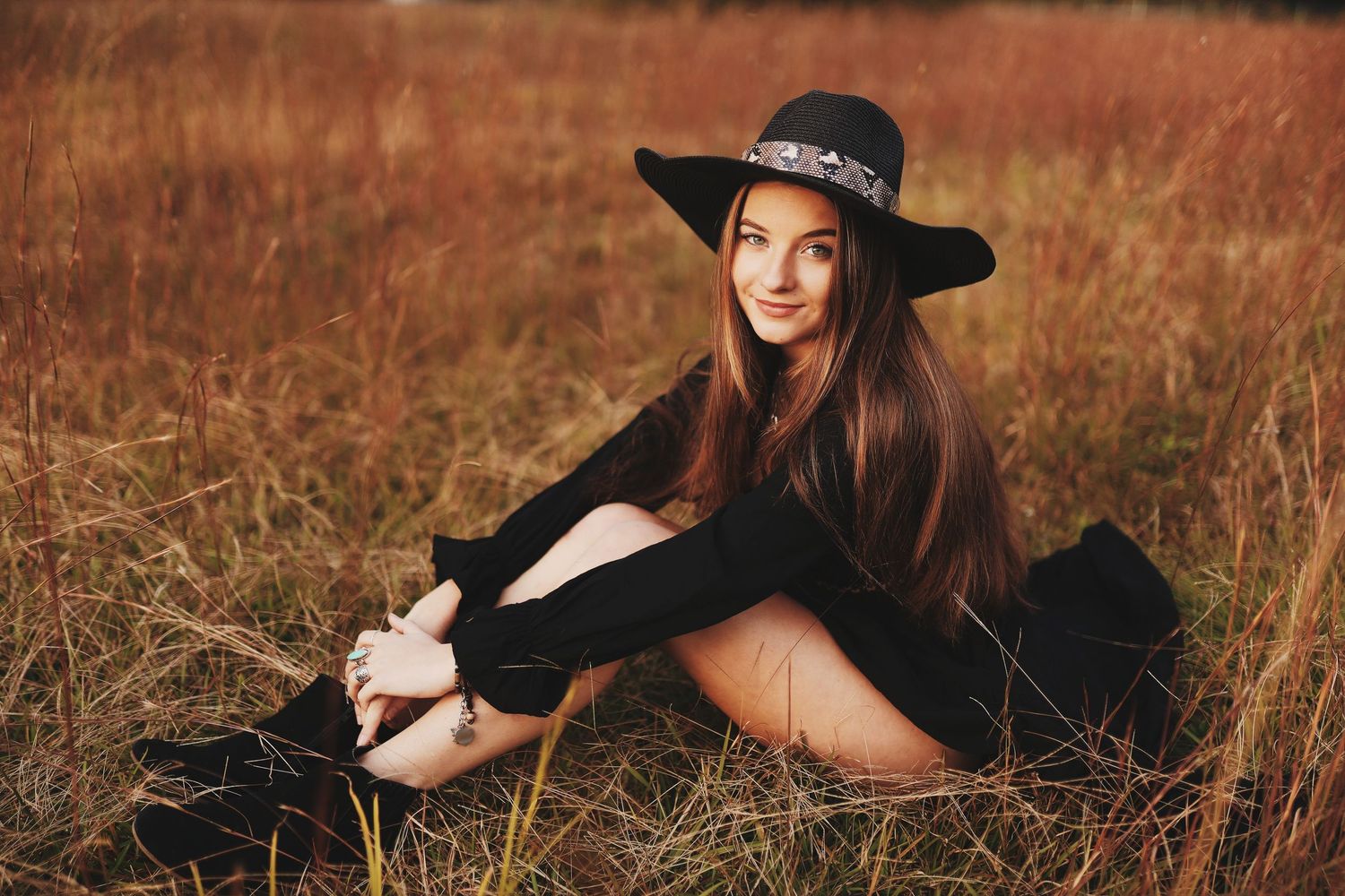 A person in a black hat and dress poses gracefully in a golden autumn field at sunset.