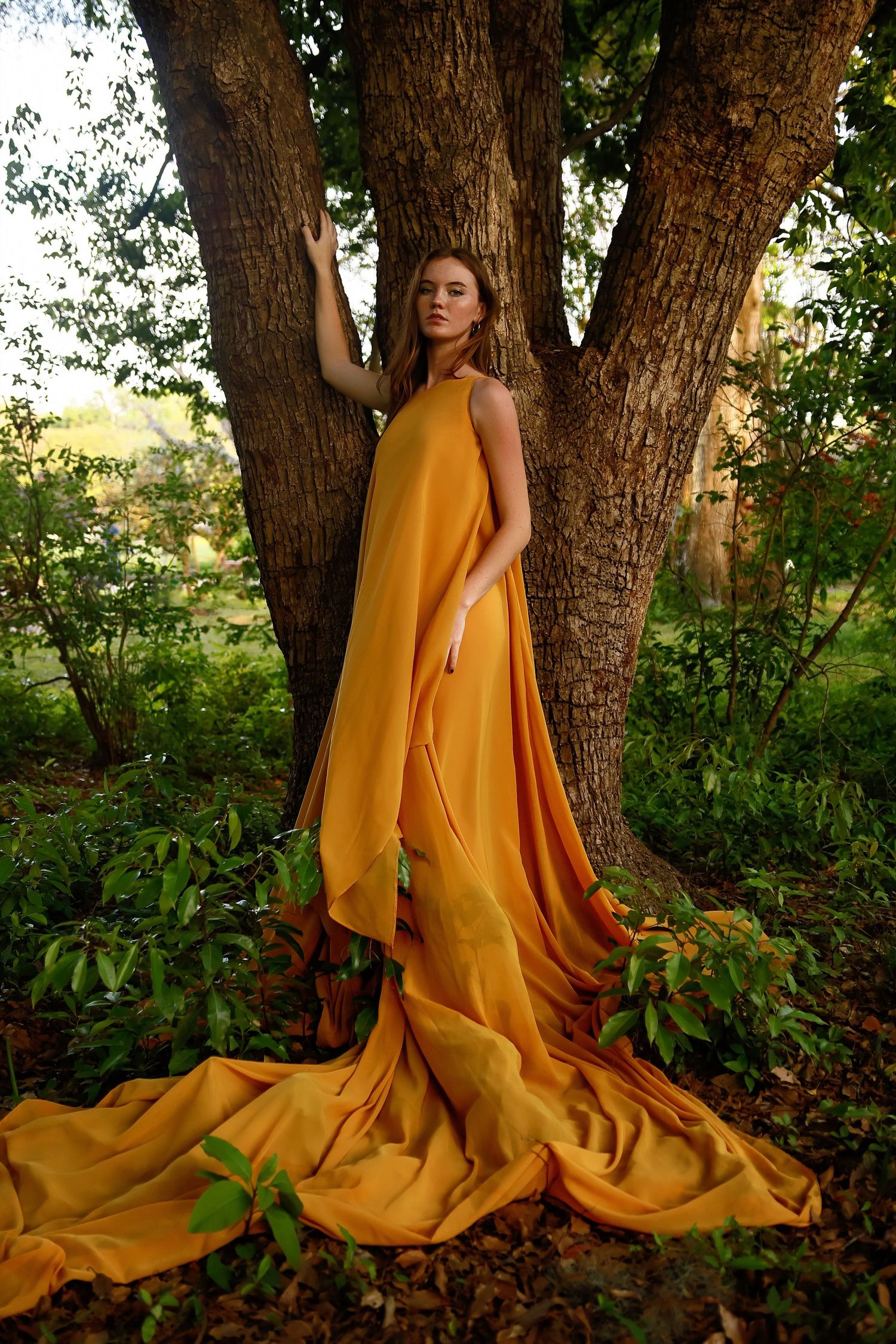 Flowing golden dress cascades down against natural forest backdrop with sunlight filtering through trees.
