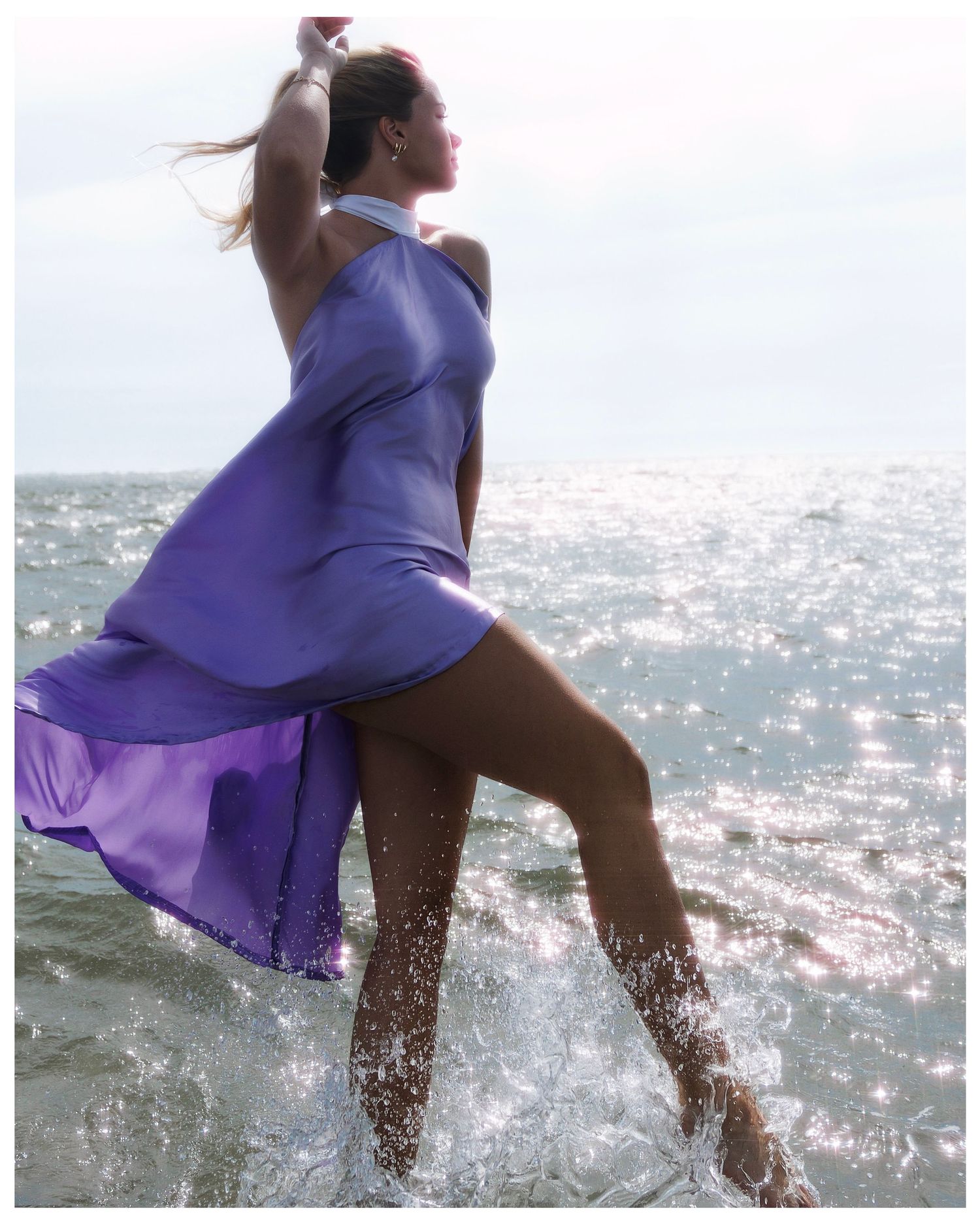 A dancer in a flowing purple dress leaps gracefully through sparkling ocean waves on a sunny day.