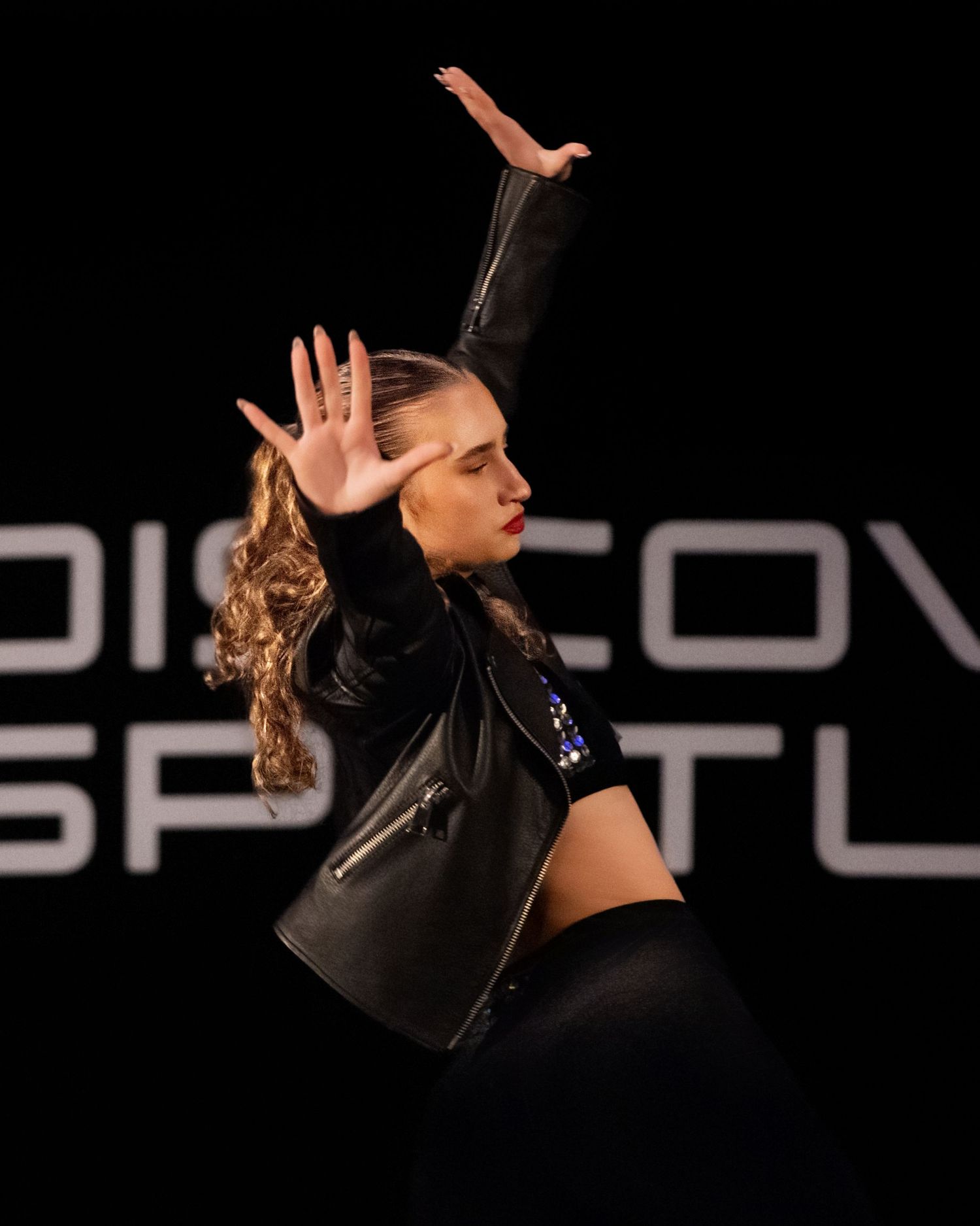 A dancer in black attire performs an expressive contemporary dance movement against a dark background.