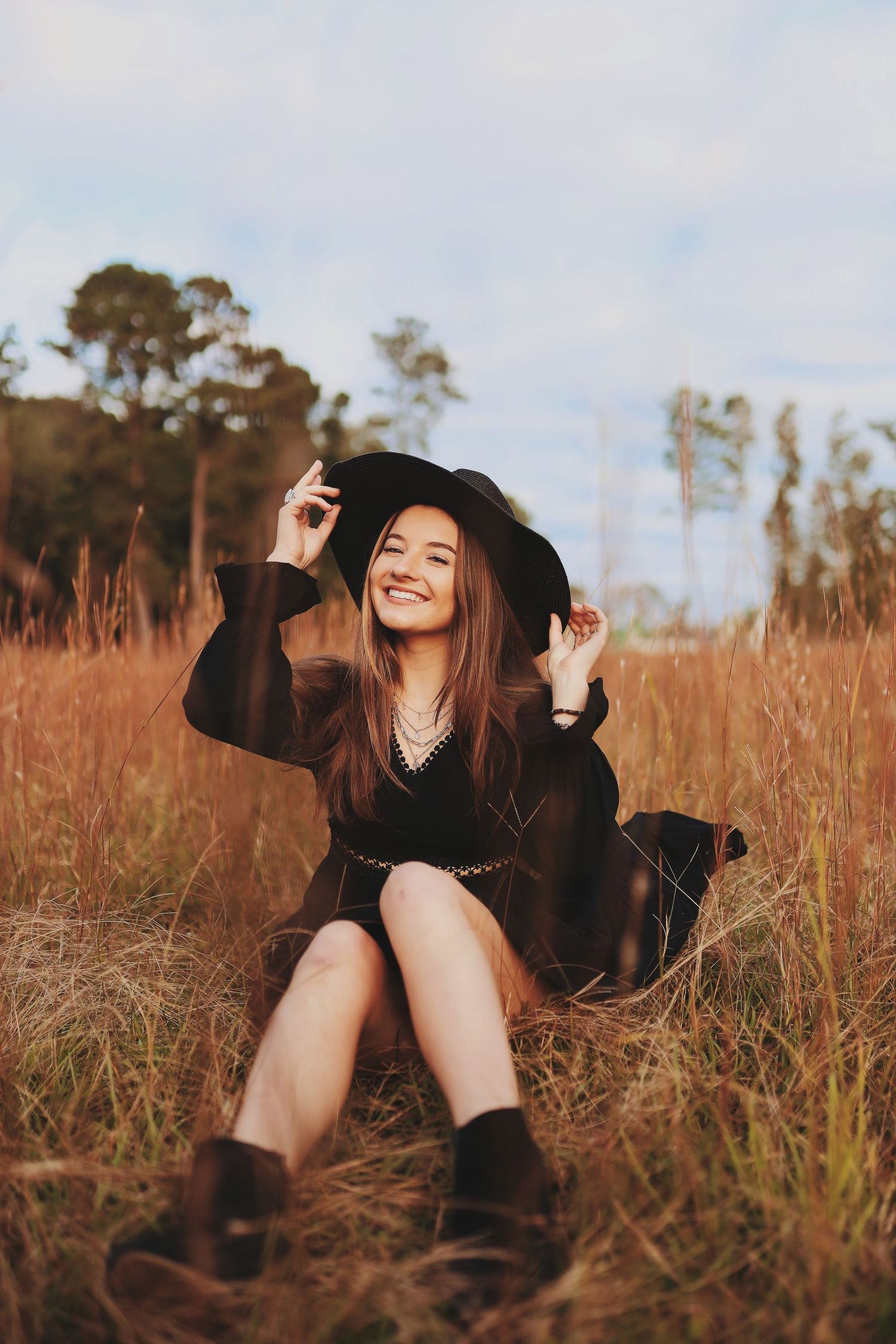 A person in a black outfit lies playfully in tall golden grass during sunset.