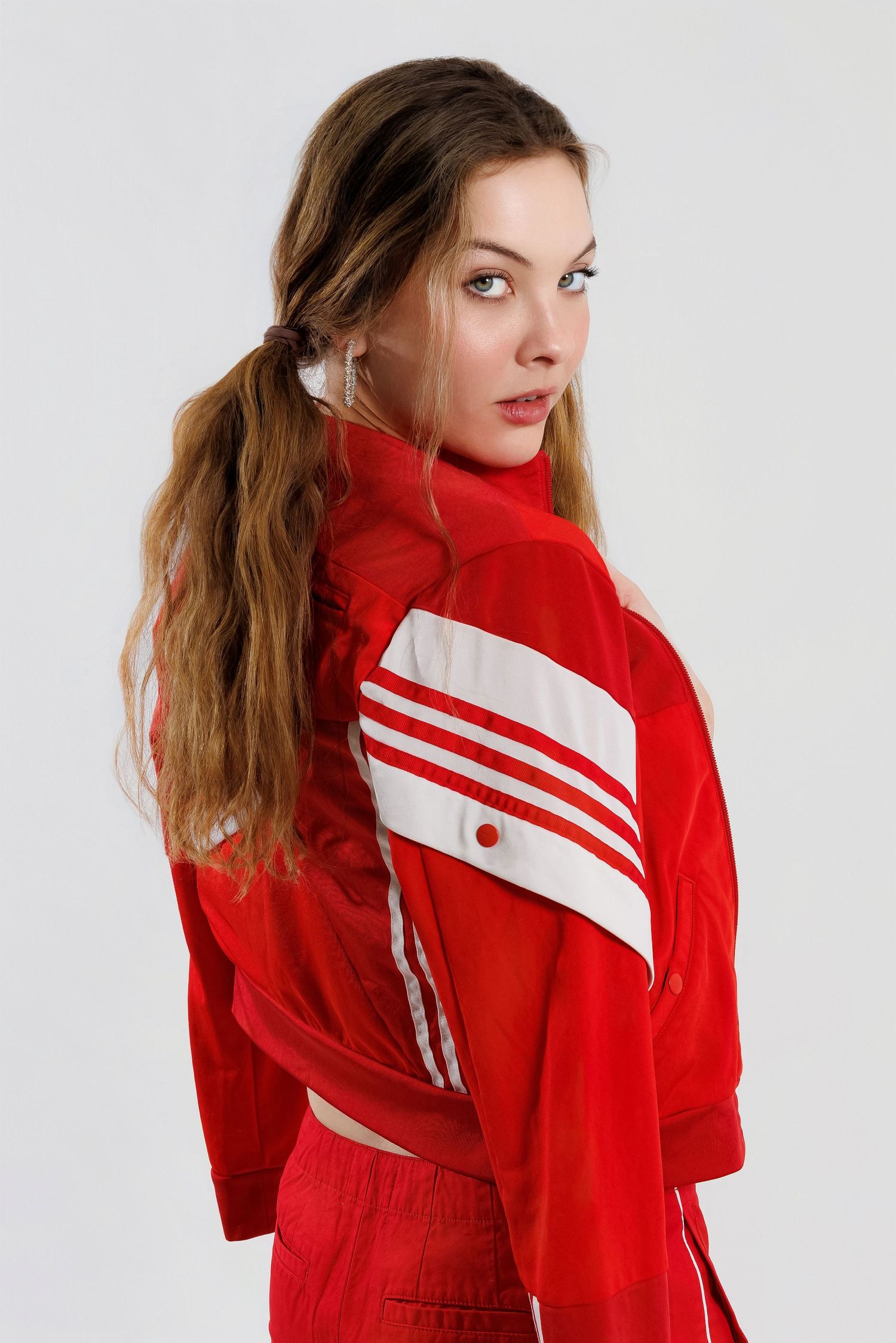 A person in a bright red track jacket with white stripes poses against a white background looking over their shoulder.