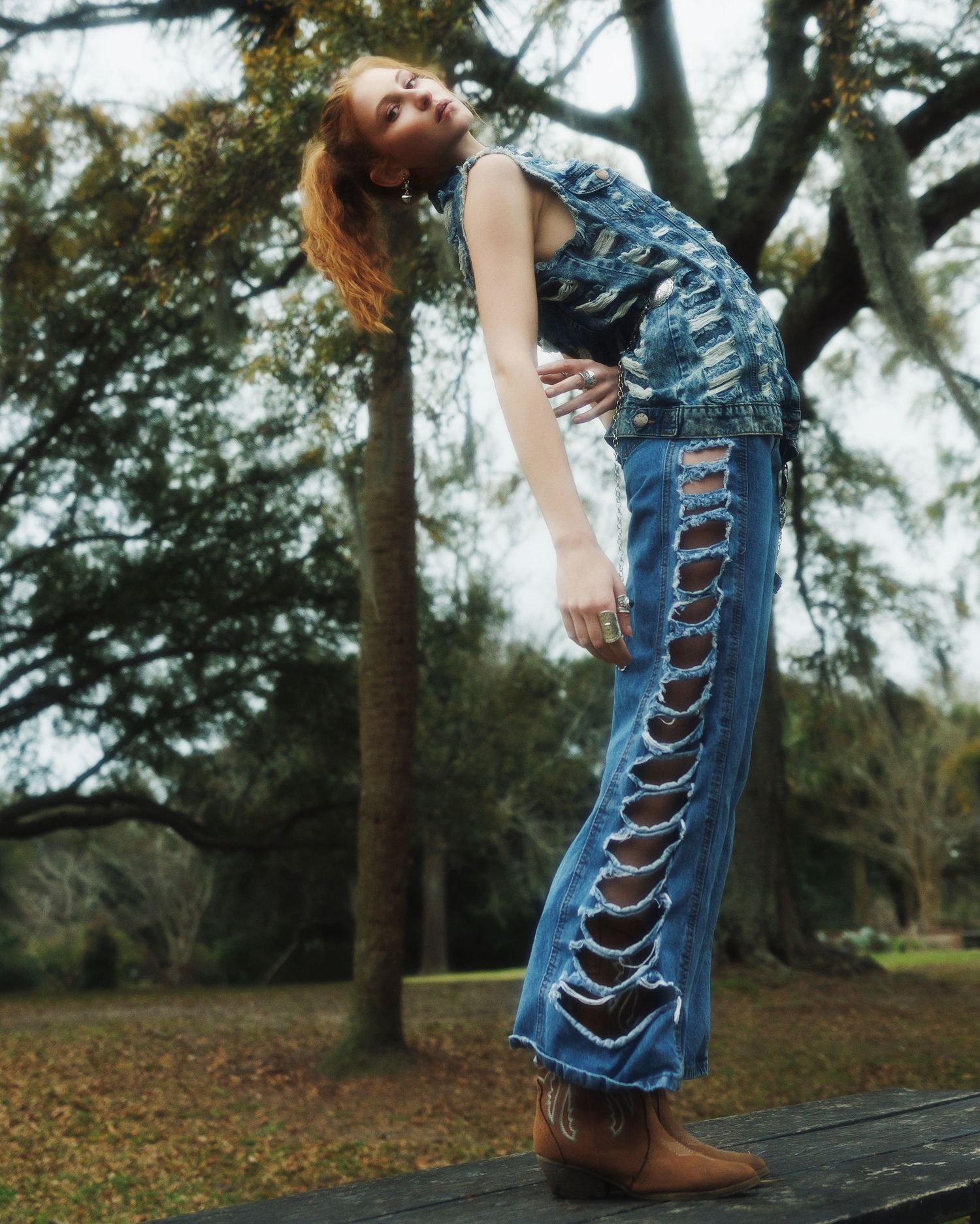 Person in retro-style distressed denim overalls poses gracefully against trees in a moody outdoor setting.