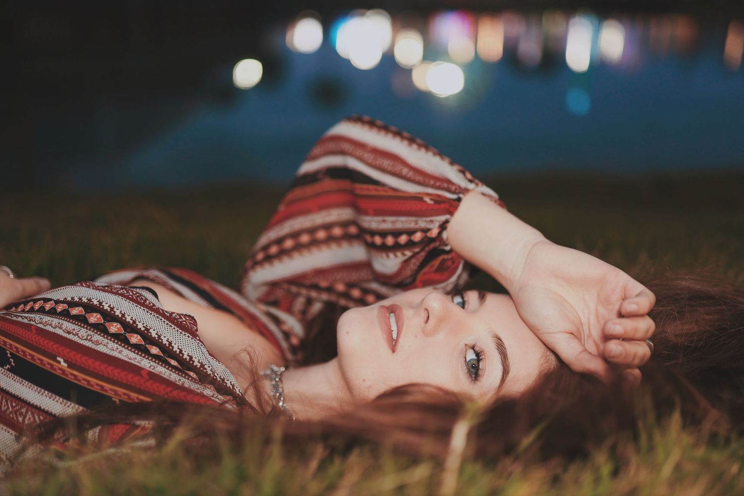 A woman in a red dress lies dreamily in a field of tall grass at sunset with bokeh lights in the background.