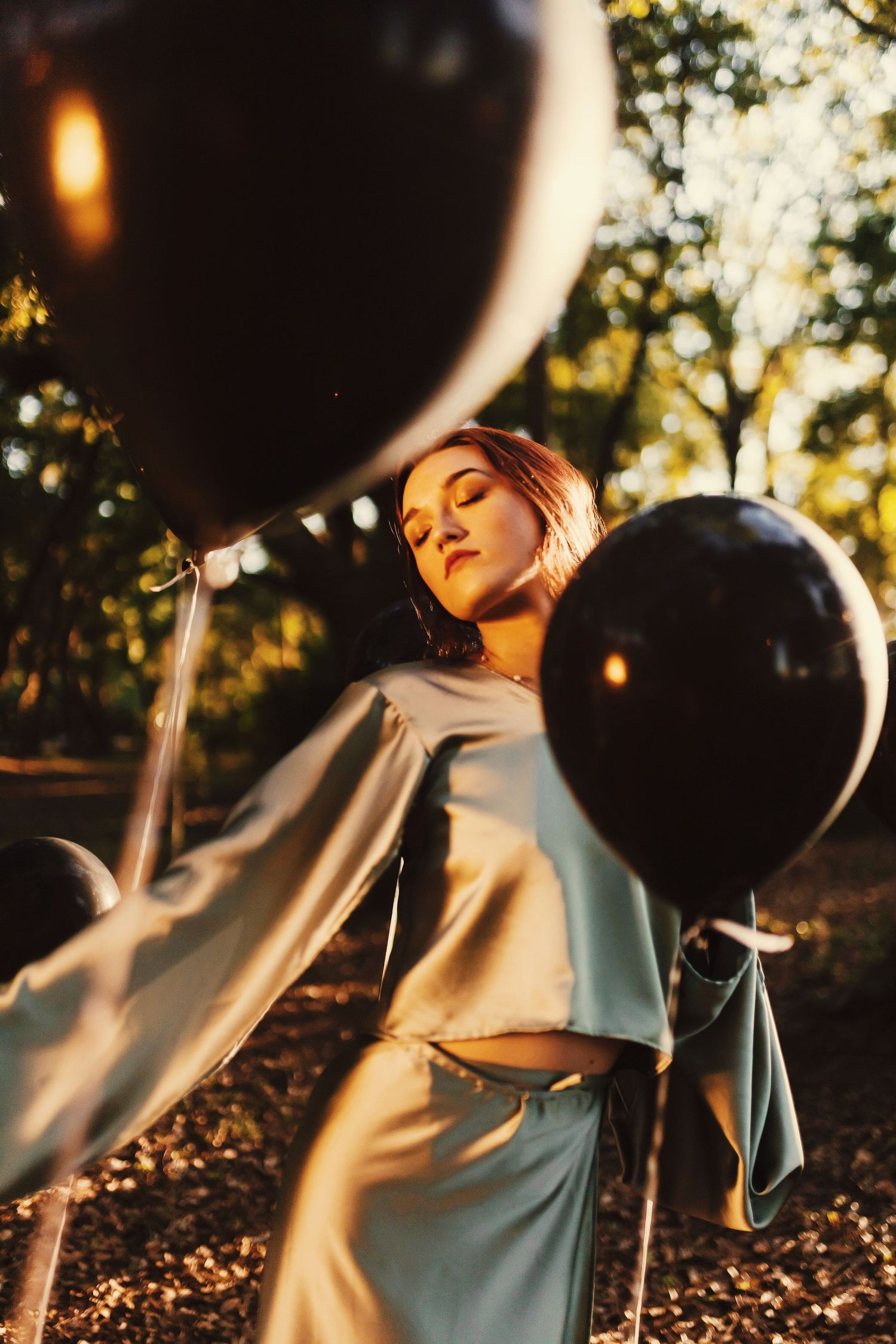 Person in light blue top poses outdoors with black balloons during golden hour sunset lighting.
