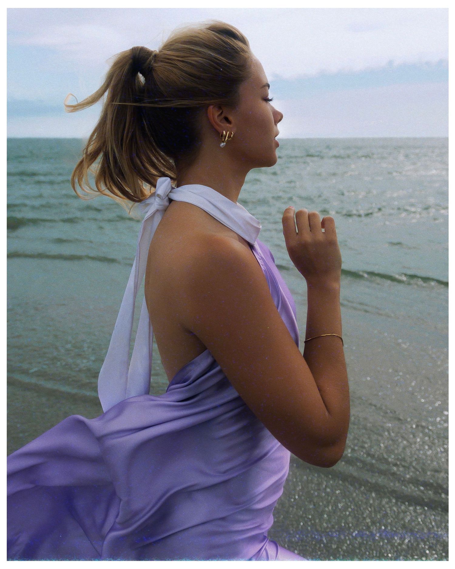 Someone wearing a lavender tank top stands thoughtfully at the ocean's edge during sunset.