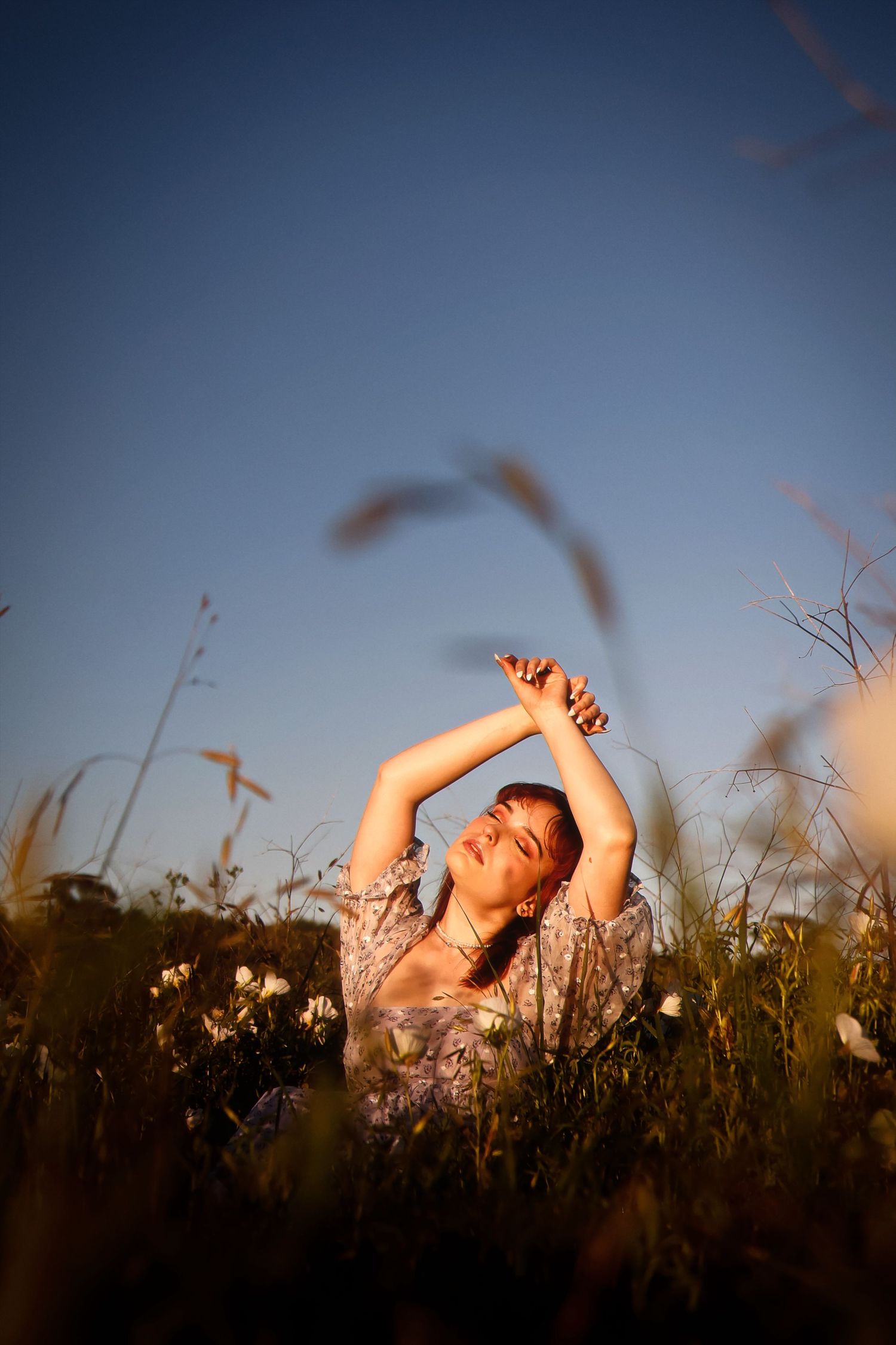 A person lies in a golden field of tall grass at sunset with arms raised overhead against a dusky blue sky.