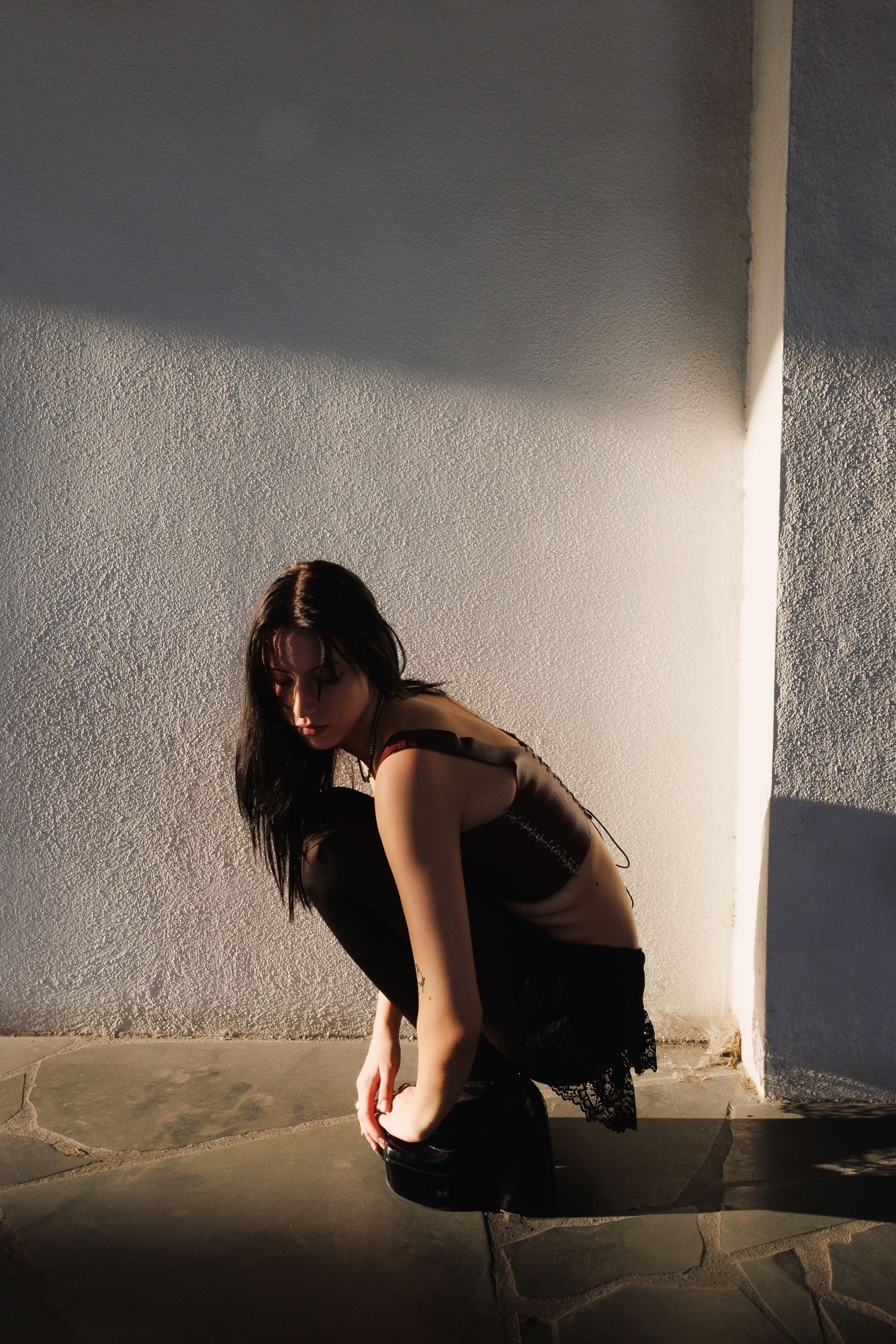 A figure crouches in shadow against a textured white wall, with dramatic side lighting creating a moody atmosphere.