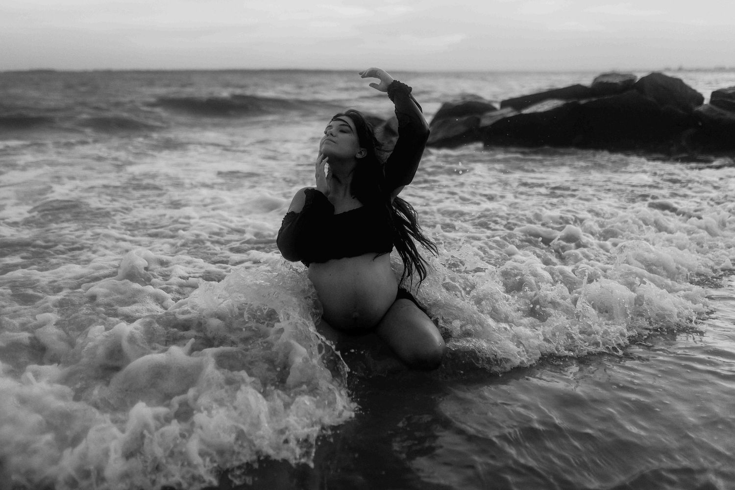 Black and white photo of a woman in bikini sitting in ocean waves near rocks, hair flowing, dramatic seascape.