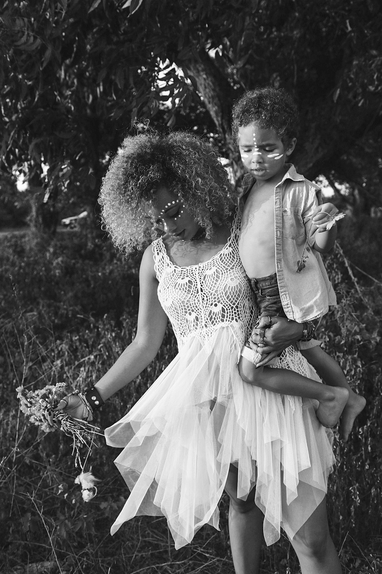 Black and white photo of a woman in white dress holding a child with face paint outdoors.