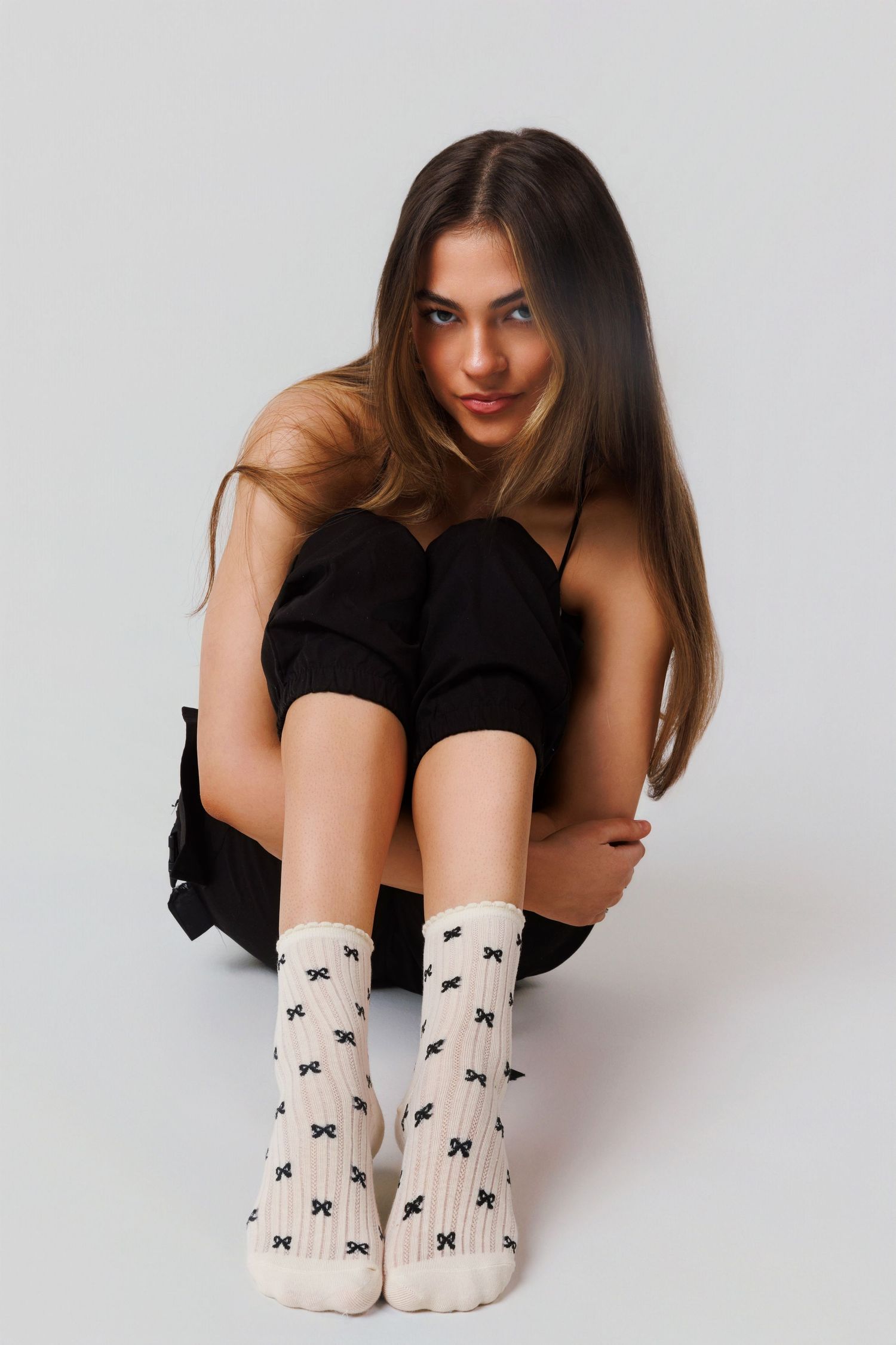 Model in casual black outfit sits on floor wearing white socks with bow pattern against plain gray background.