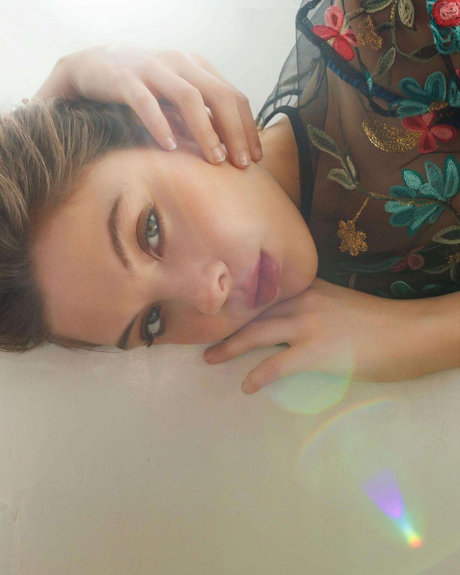 Close-up portrait showing someone wearing a floral embroidered sheer top with a rainbow light effect in frame.