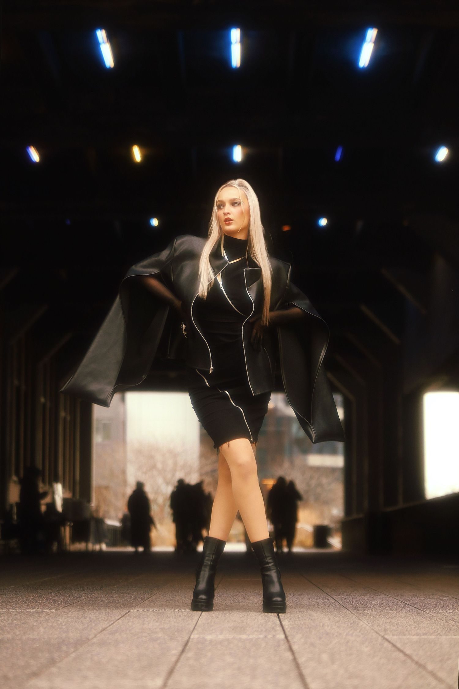 Model in black dress and leather boots poses under dramatic lighting on concrete walkway with silhouetted figures behind.