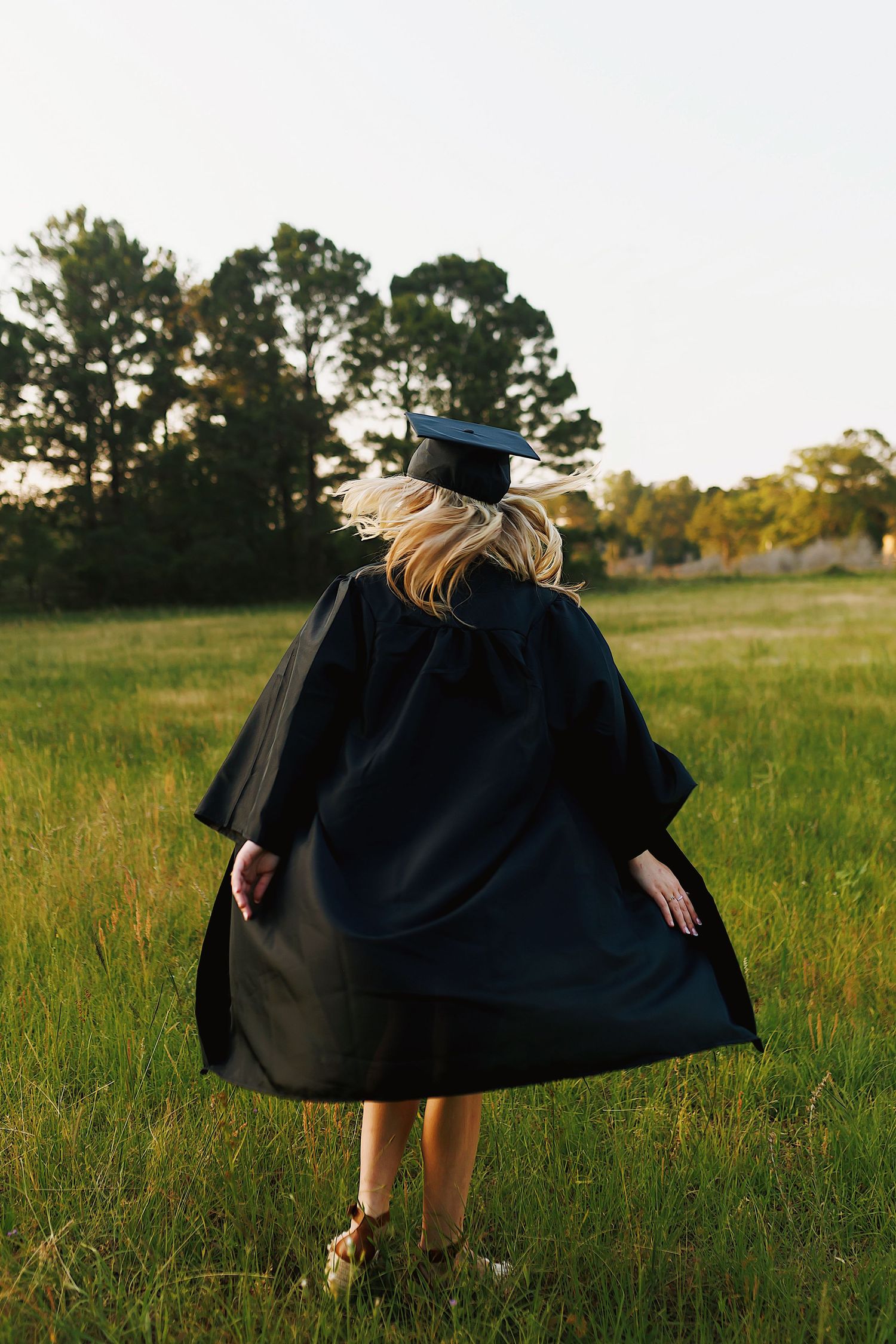 Graduate in black cap and gown walking through a sunlit field at golden hour.