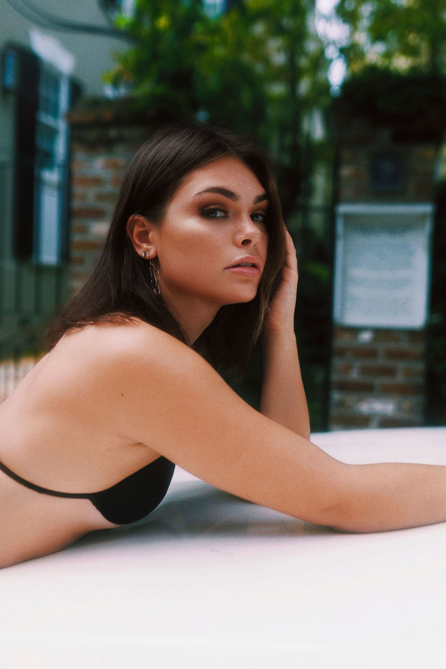 A person in a black top rests on a white surface in an outdoor setting with brick walls and greenery behind them.