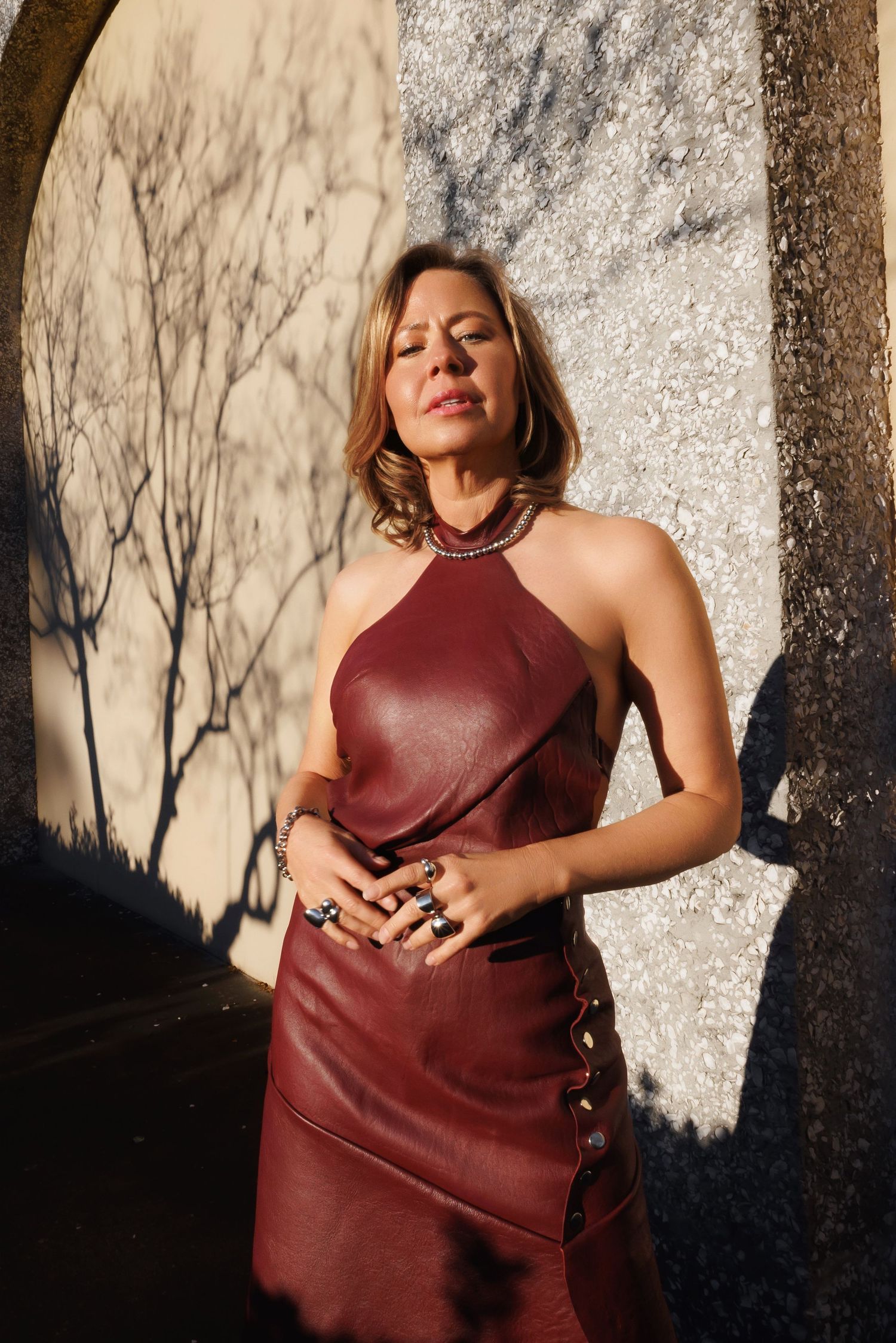Person in burgundy leather halter dress poses against textured wall with dramatic shadows and natural lighting.