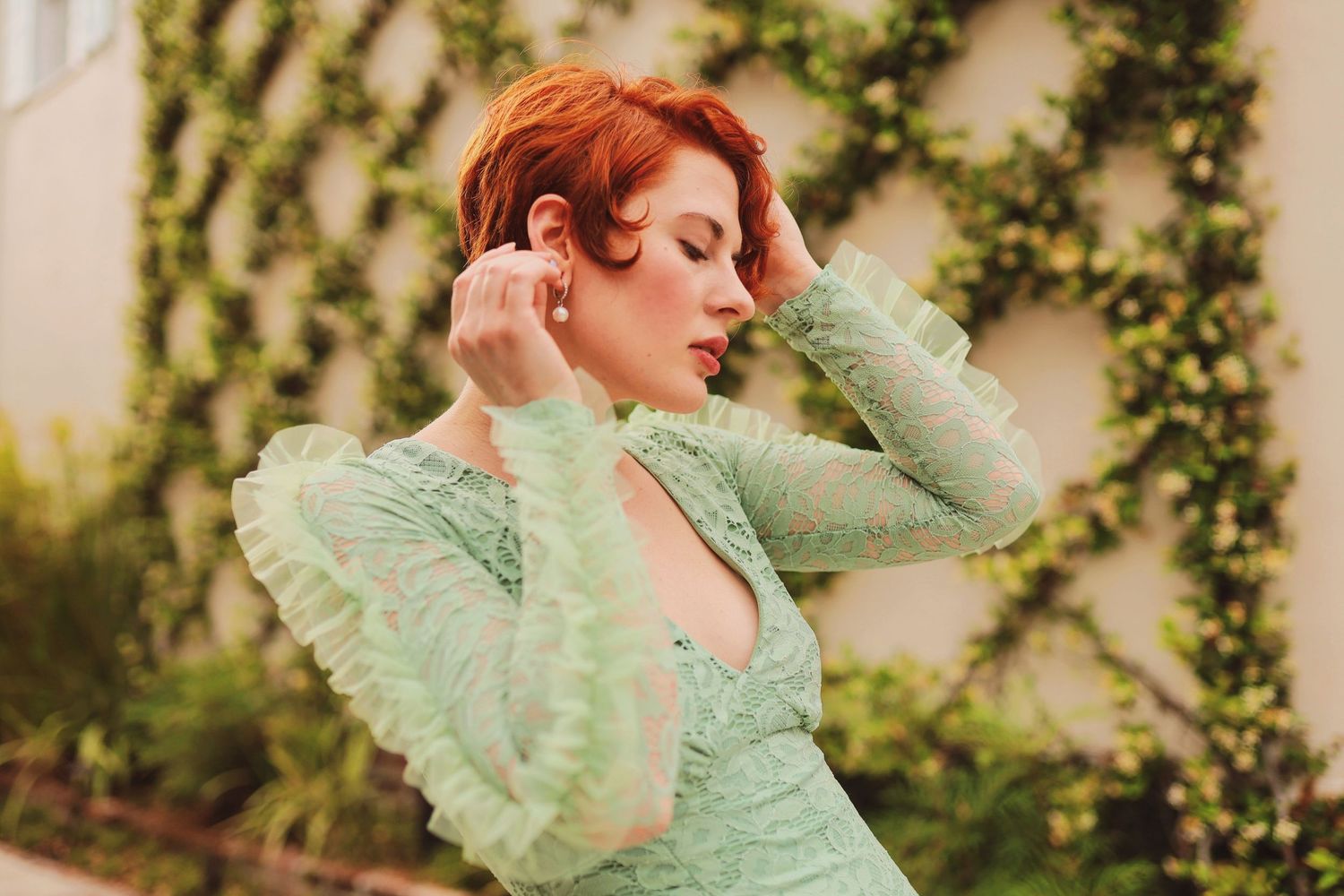 Woman in mint green ruffled dress poses gracefully against a lush garden backdrop with climbing vines.