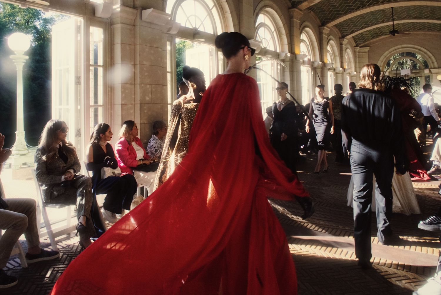 A person in a flowing red dress walks through an ornate arched hallway with natural light streaming through windows.