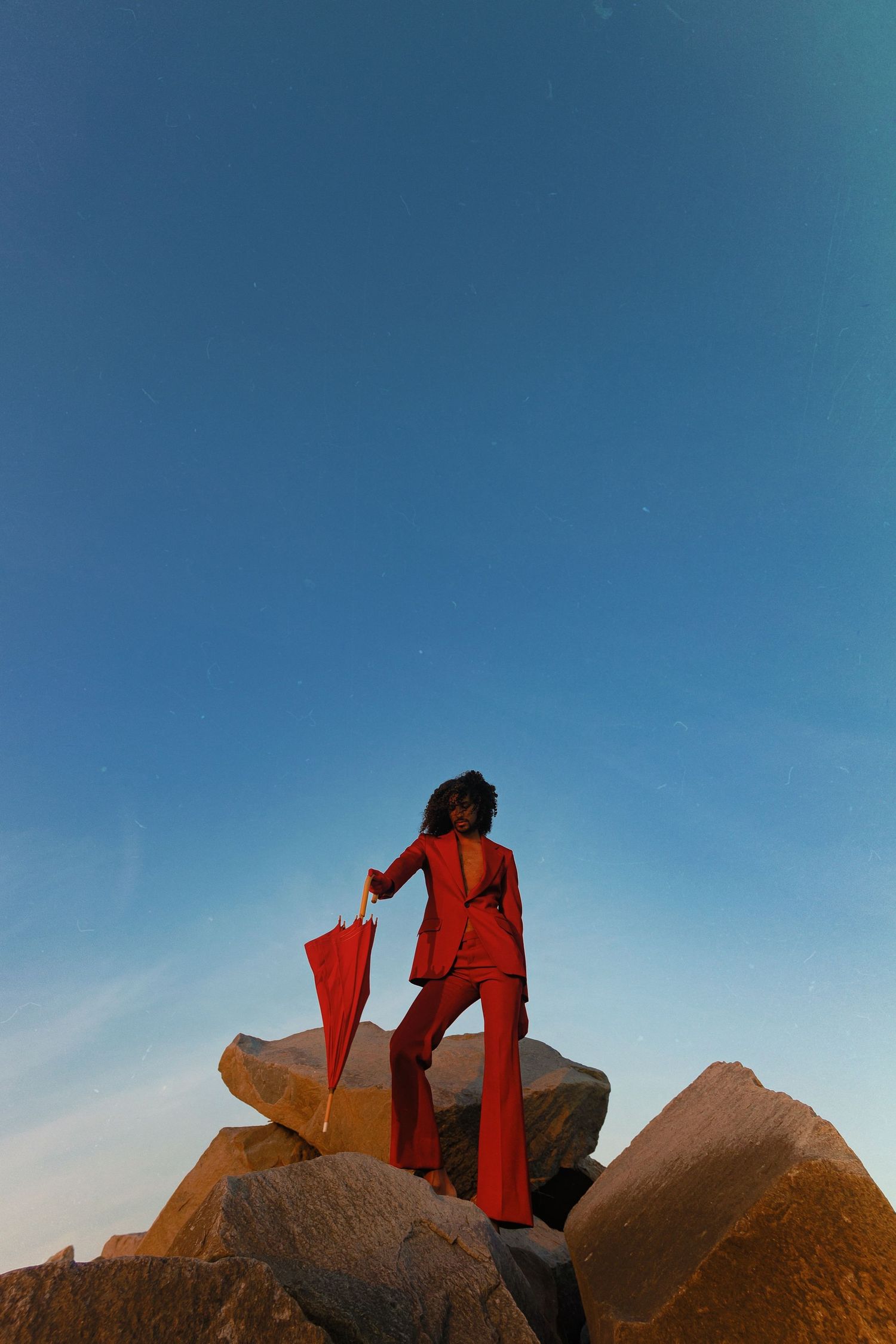 A figure in a red outfit is silhouetted against a bright blue sky while resting on a rock formation.