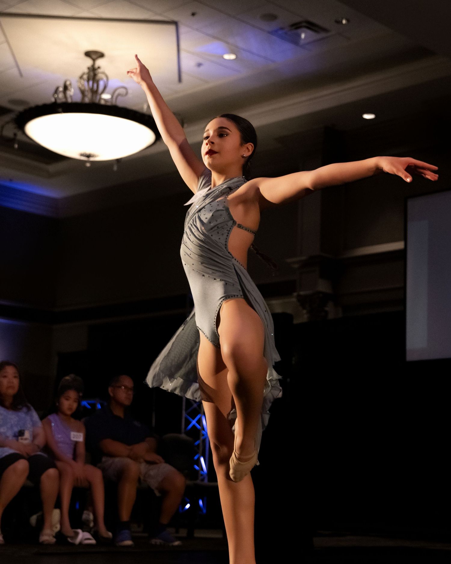 A dancer performs an elegant leap in a dimly lit venue with dramatic lighting and audience silhouettes visible.