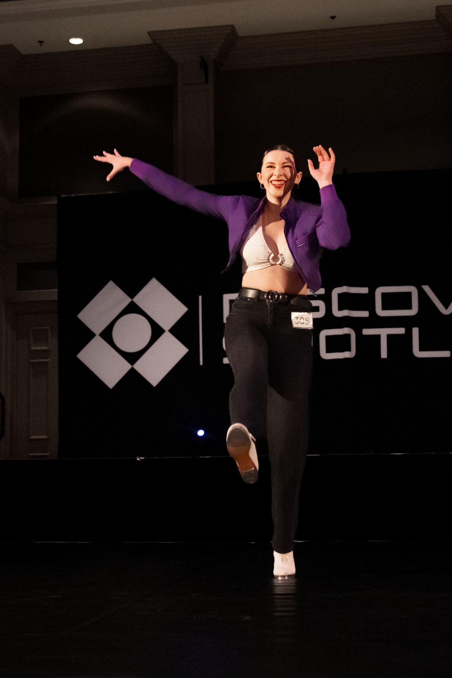 A dancer in black leggings and purple top performs dynamic movements in a dimly lit studio with a logo on the wall.