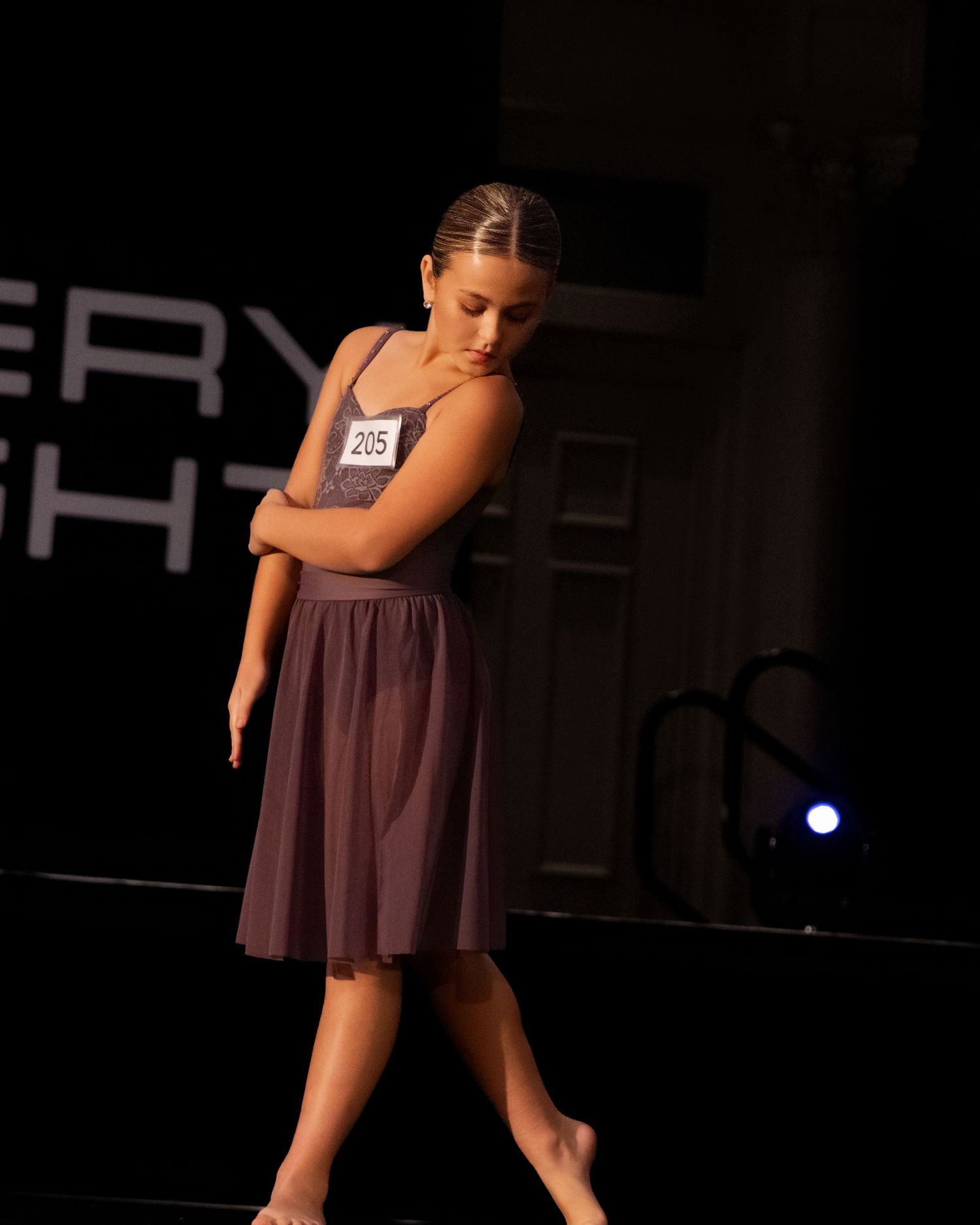A dancer in a flowing brown dress performs an expressive contemporary dance movement on a dark stage.