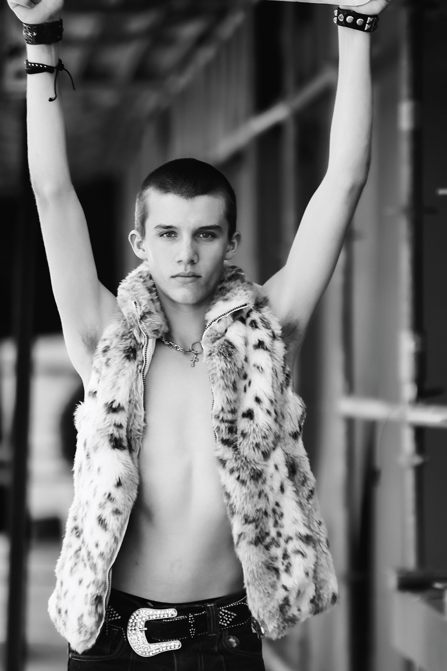 A person in a spotted fur vest poses with raised arms against a blurred background in black and white.