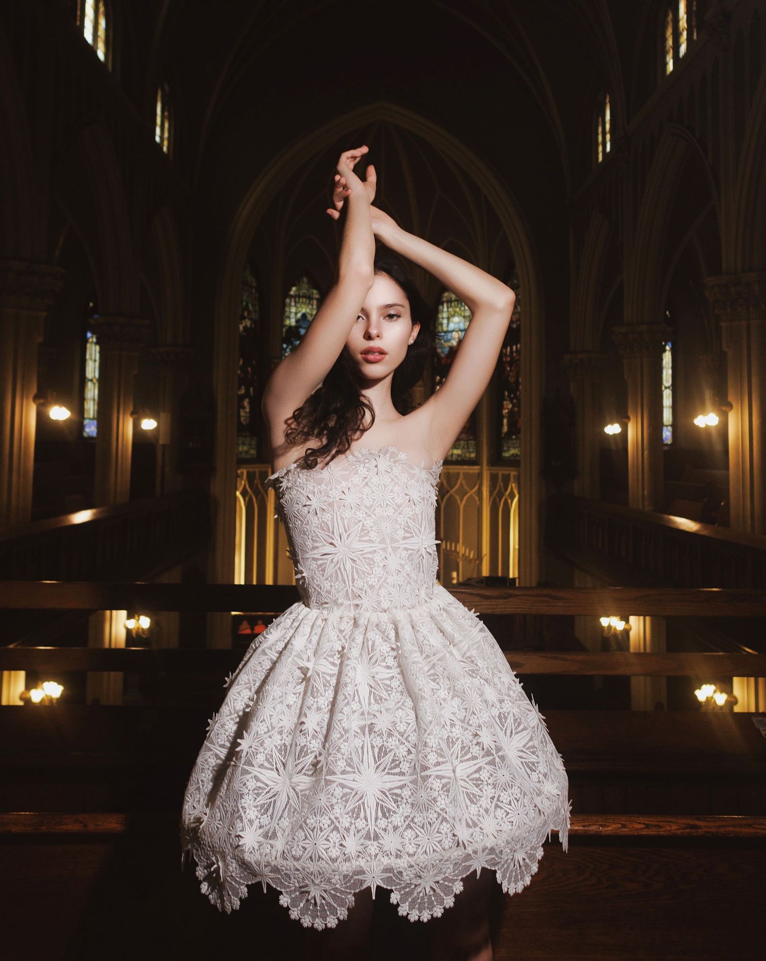 A figure in a white lace dress poses dramatically in a dimly lit gothic architectural space with arched windows and columns.
