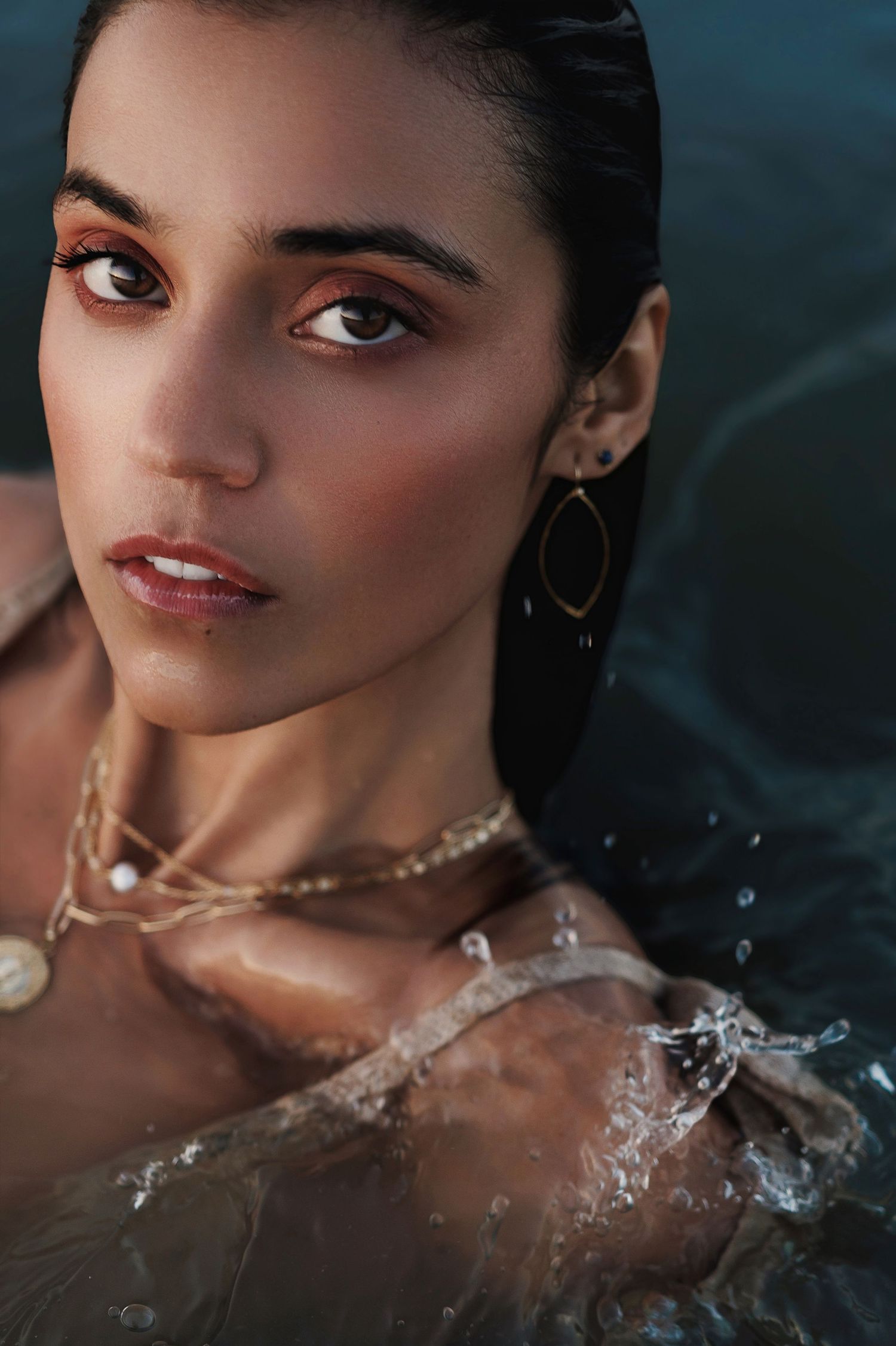 Close-up beauty portrait with delicate gold jewelry in water, featuring warm tones and artistic lighting.