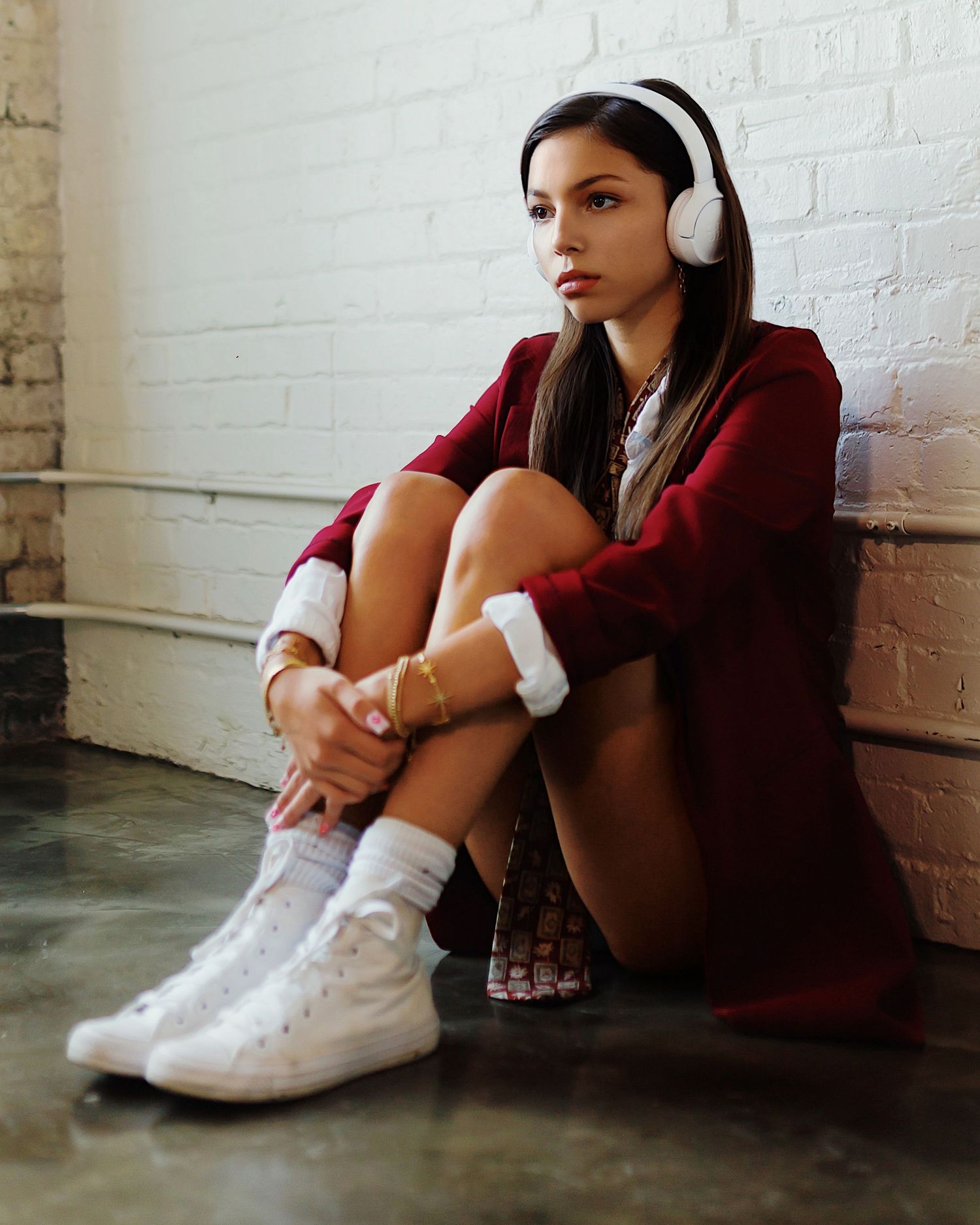 Someone wearing white headphones, burgundy sweater and white sneakers sits on floor against white painted brick wall.