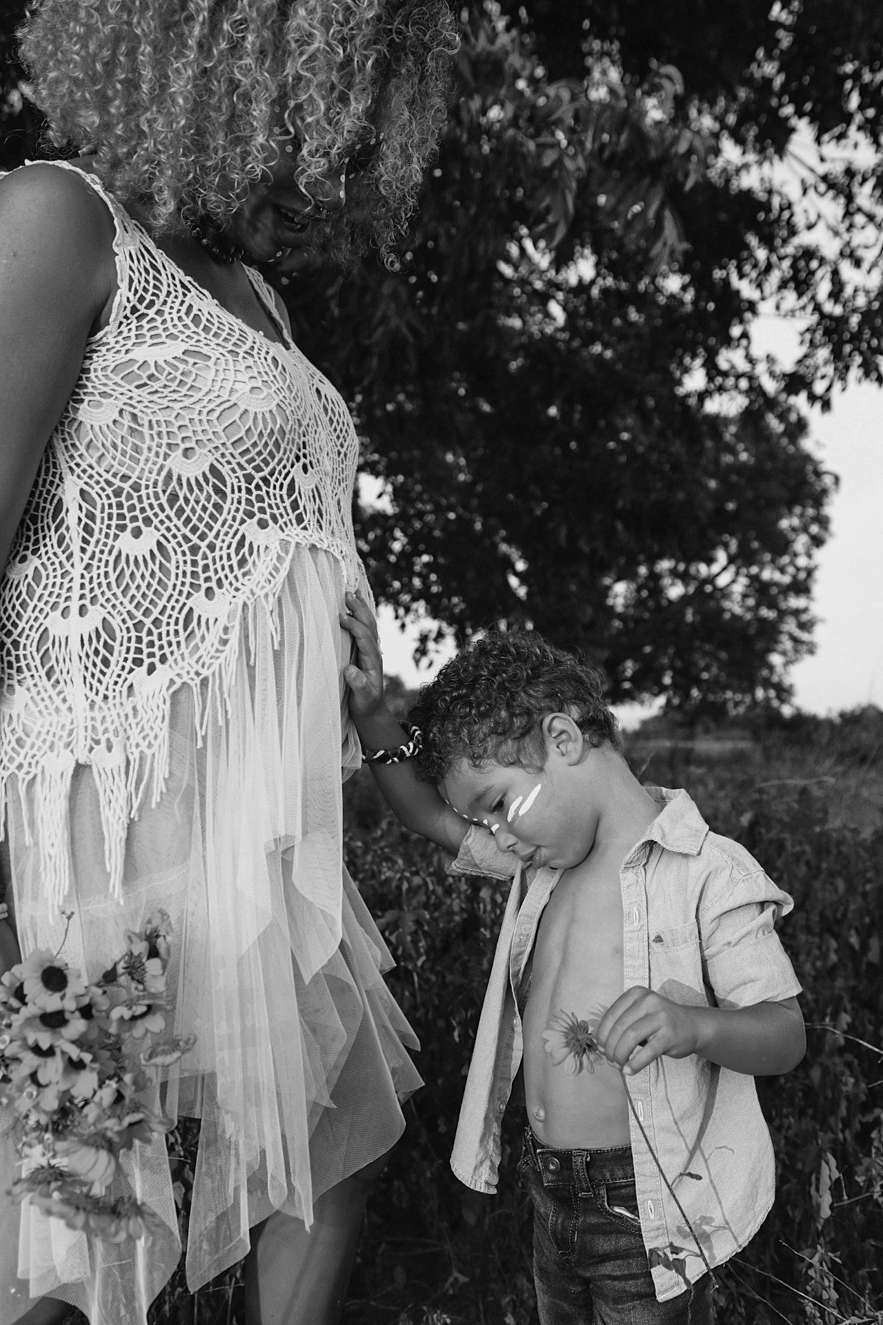 Black and white photo of a young boy standing beside a pregnant woman in a lace dress outdoors.