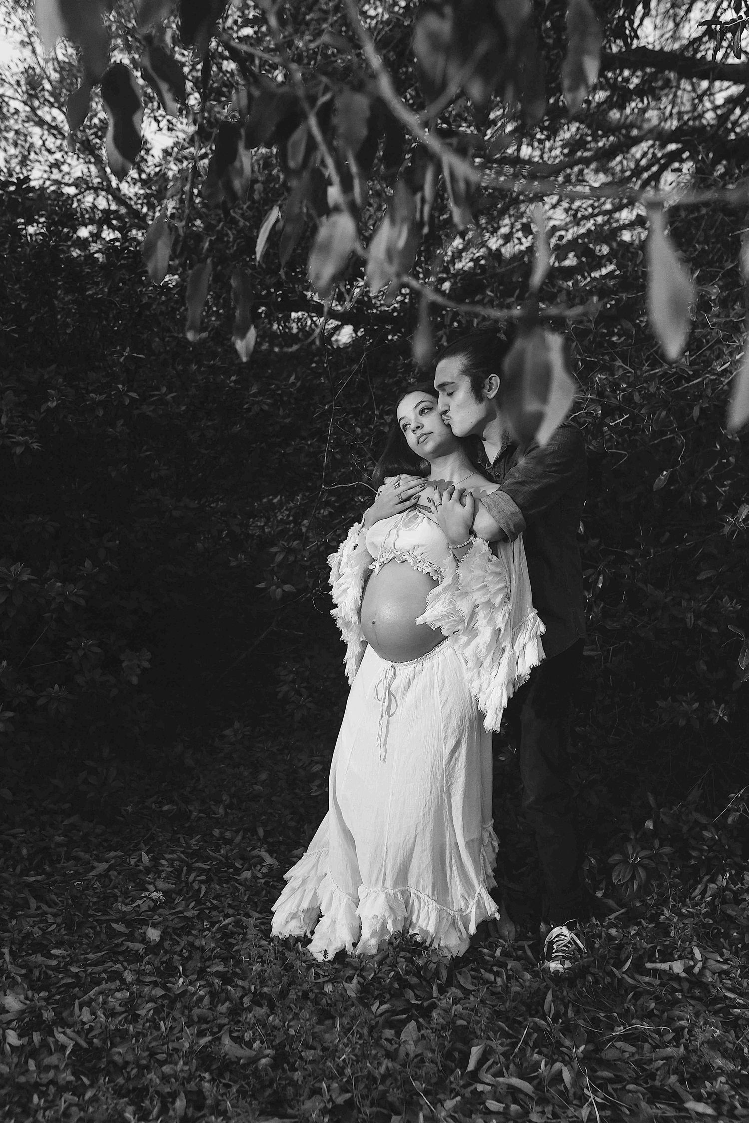 Black and white maternity photo of a couple embracing outdoors, man kissing pregnant woman in lace dress among lush foliage.