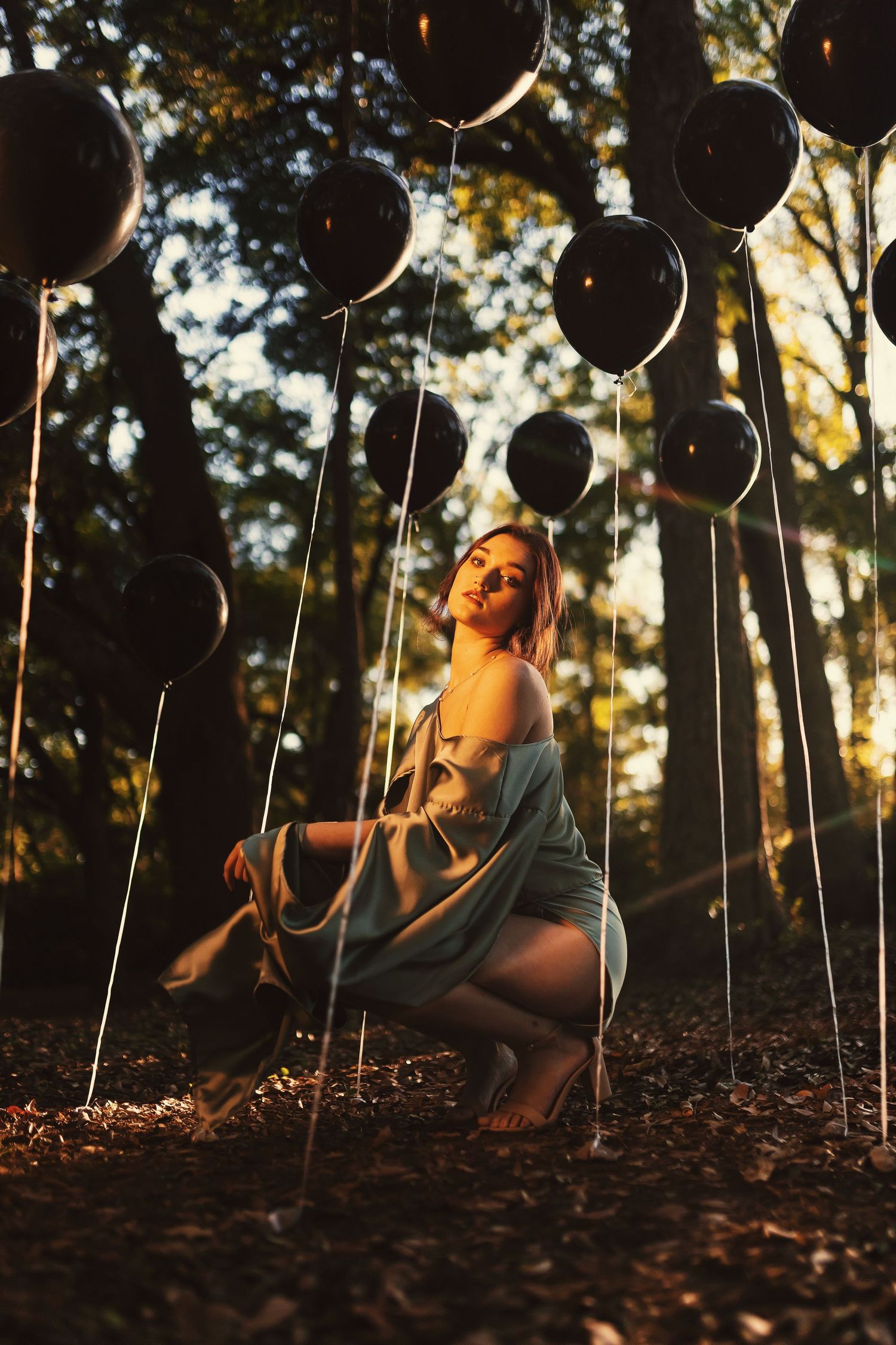A series of black balloons float among trees at sunset while someone sits on fallen leaves in warm golden light.