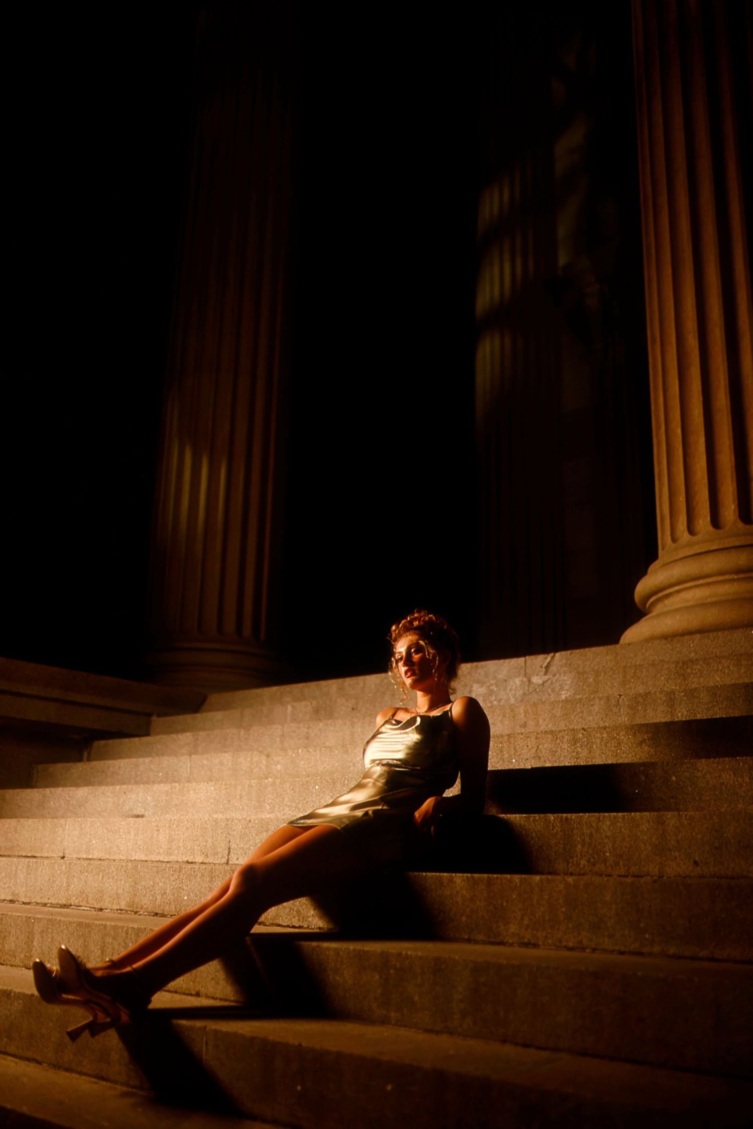 Dramatic night portrait on stone steps between illuminated classical columns with warm lighting creating shadows.