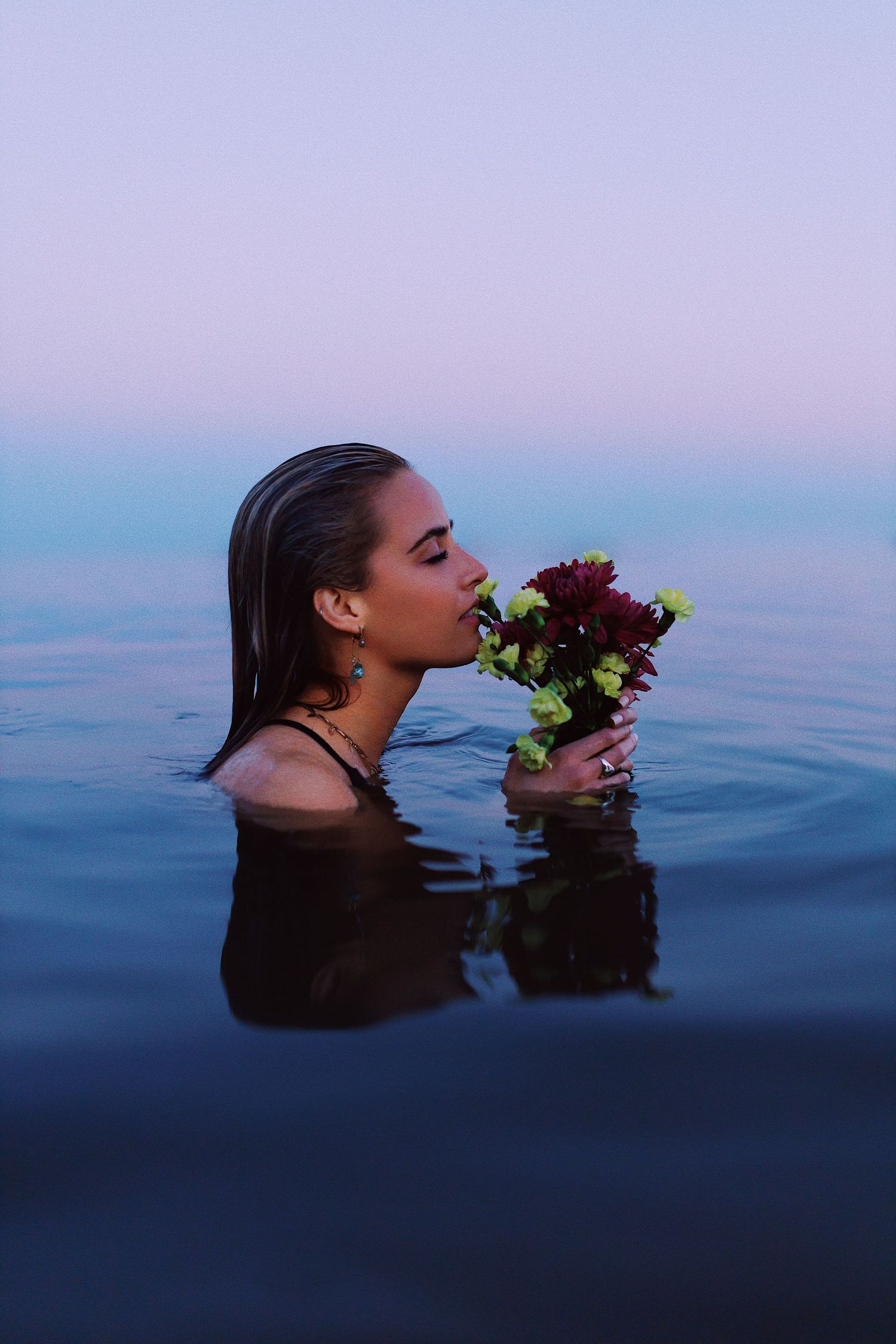 A figure emerges from dark blue water holding a bouquet of burgundy flowers against a dreamy twilight sky.