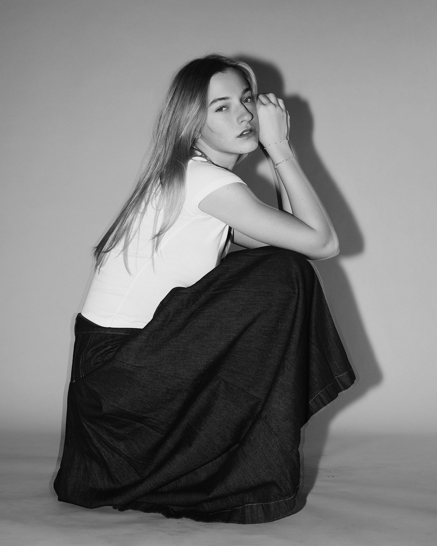 A black and white portrait of a figure in a white top and black skirt crouched against a plain background.